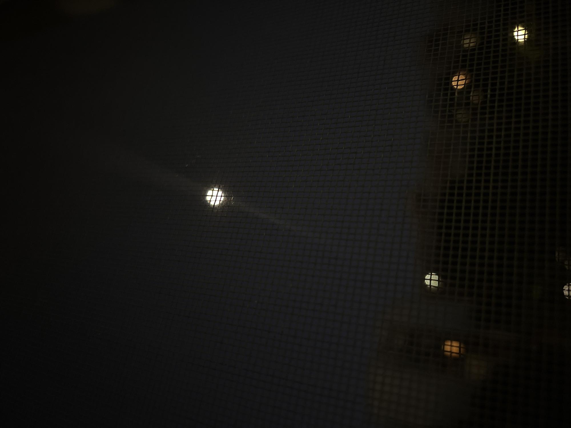 The image appears to be taken from indoors, looking out through a window screen. The view is of an urban scene at night. Multiple lights, possibly from buildings across from the window, are visible through the screen. The grid pattern of the screen is superimposed on the lights. The scene's darkness suggests it is night. It is impossible to determine the exact location in Plovdiv, Bulgaria.