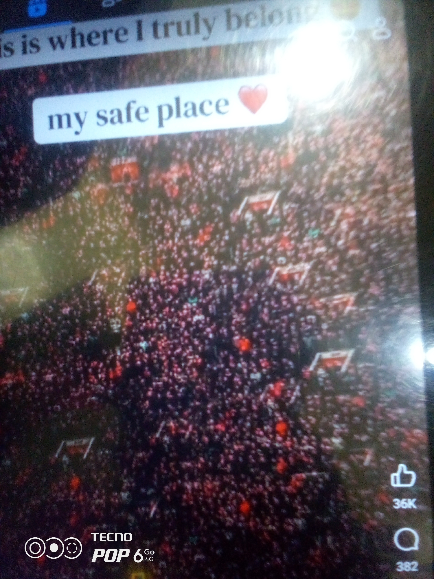 The image displays a screen with a video overlayed with text. The top text reads "is where I truly belong". Below that, a white oval with rounded corners contains the text "my safe place" followed by a red heart icon. The background of the screen shows a large crowd of people, mostly wearing dark clothing, with some red and green accents visible. Several flags with red and white designs are held up in the crowd. The bottom of the screen shows the branding "TECNO POP 6 4G" and icons for likes and comments with counts of "36K" and "382" respectively. There is no information in the image to suggest the location is in the city of Howayi, Nigeria.