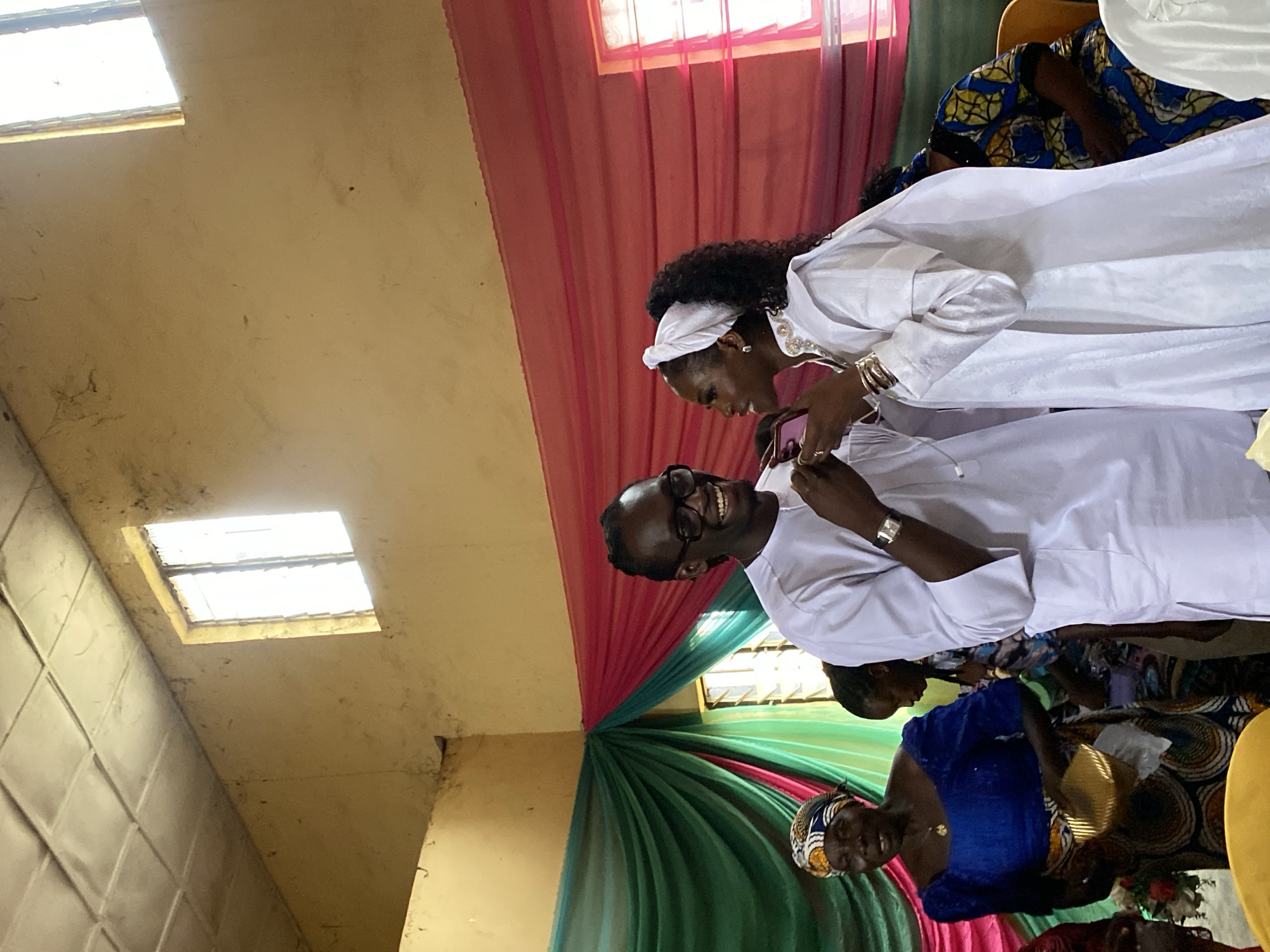 In Riyom, Nigeria, an indoor event is underway. A man with glasses and a white tunic smiles broadly, looking at a woman beside him. She is also wearing white, with a headscarf and jewelry, and appears to be holding a phone. In the background, a woman in a blue dress and headscarf holds a golden object, and other people are visible. The room has a tiled ceiling and yellow walls, with green and pink draped fabric decorating the space, possibly indicating a celebration or ceremony. Sunlight streams through a window.