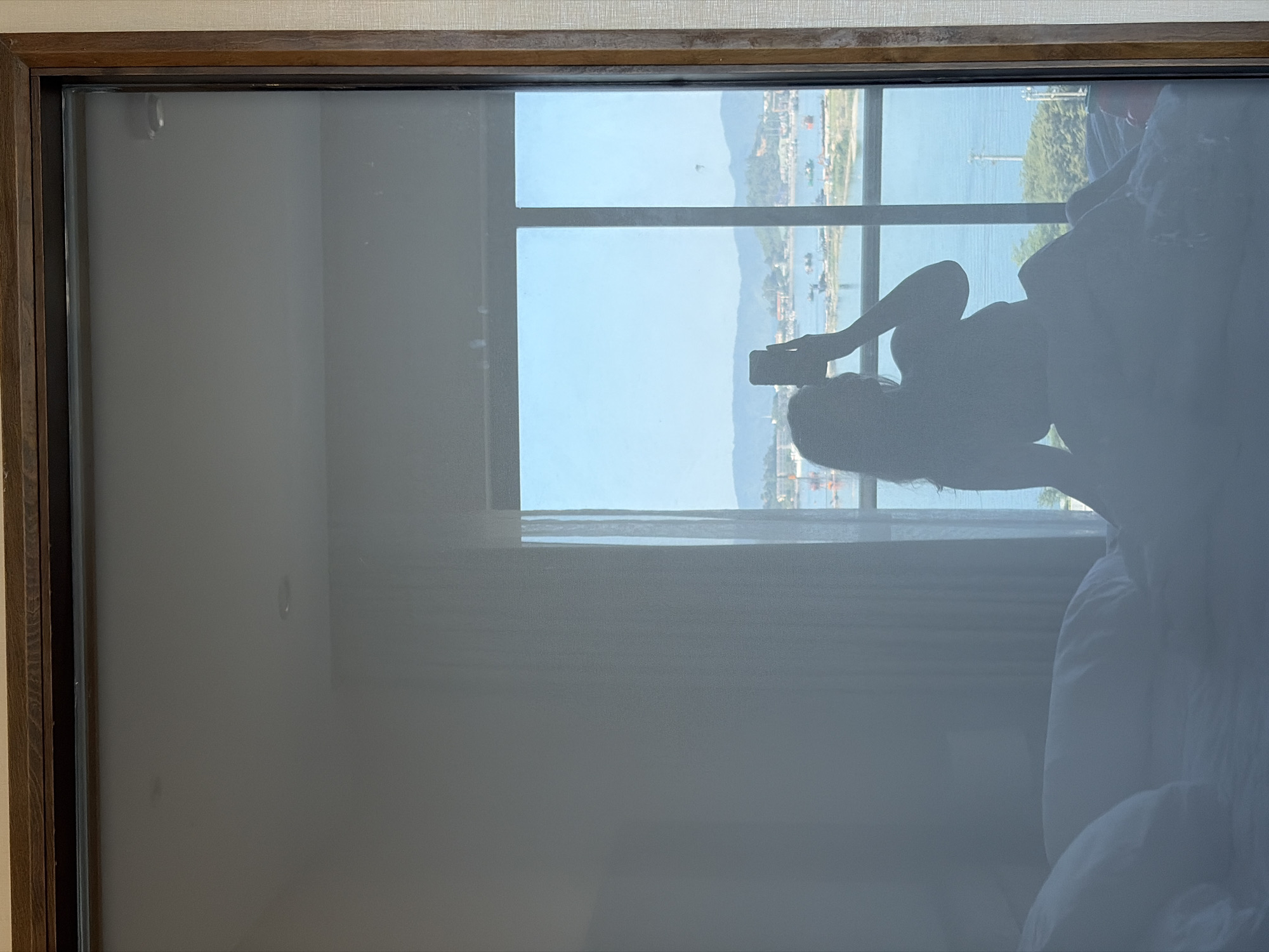 The image displays a reflection in a large window, vertically oriented and rotated 90 degrees counter-clockwise. A prominent silhouette of a person, likely female, occupies the right side of the frame, holding a smartphone to take a photo. This suggests the person is capturing their reflection or a selfie against the backdrop of the outdoor view.
Through the window, a bright, sunlit outdoor scene is visible under a clear sky. This view features a large body of water, identifiable as a bay or ocean, with distant mountains forming the horizon. Several boats or ships are scattered across the water. The architecture and landscape elements are consistent with a coastal city like Da Nang, Vietnam.
The window is divided into multiple panes by dark mullions. A sheer, light grey curtain or blind partially covers the left side of the window, extending across a significant portion of the interior space. The window is framed by a dark wooden border, with an off-white textured wall visible above it. In the lower right corner, parts of a white pillow or bedding are faintly discernible, indicating an indoor setting such as a hotel room or apartment. The bright illumination suggests it is daytime with clear weather. No text is visible in the image.