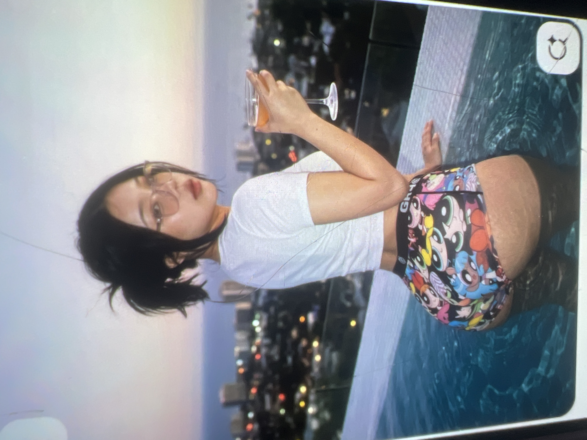 A woman with black hair pulled into a ponytail, wearing glasses, a white cropped t-shirt, and colorful shorts with cartoon characters, is standing in a swimming pool. She is holding a cocktail glass in her right hand. The background shows a city skyline at dusk with lights from buildings and the ocean. The scene appears to be on a rooftop or elevated area overlooking the city.