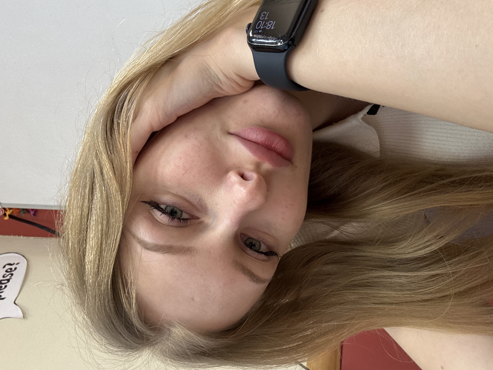 A close-up, rotated portrait depicts a fair-skinned young woman with blonde hair, wearing a dark-banded smartwatch on her left wrist displaying "18:10 13." She is looking at the camera, with her left hand partially obscuring the left side of her face. The background is out of focus but shows elements of a room, including a red wall. There is no discernible information confirming a location in Perm, Russia.