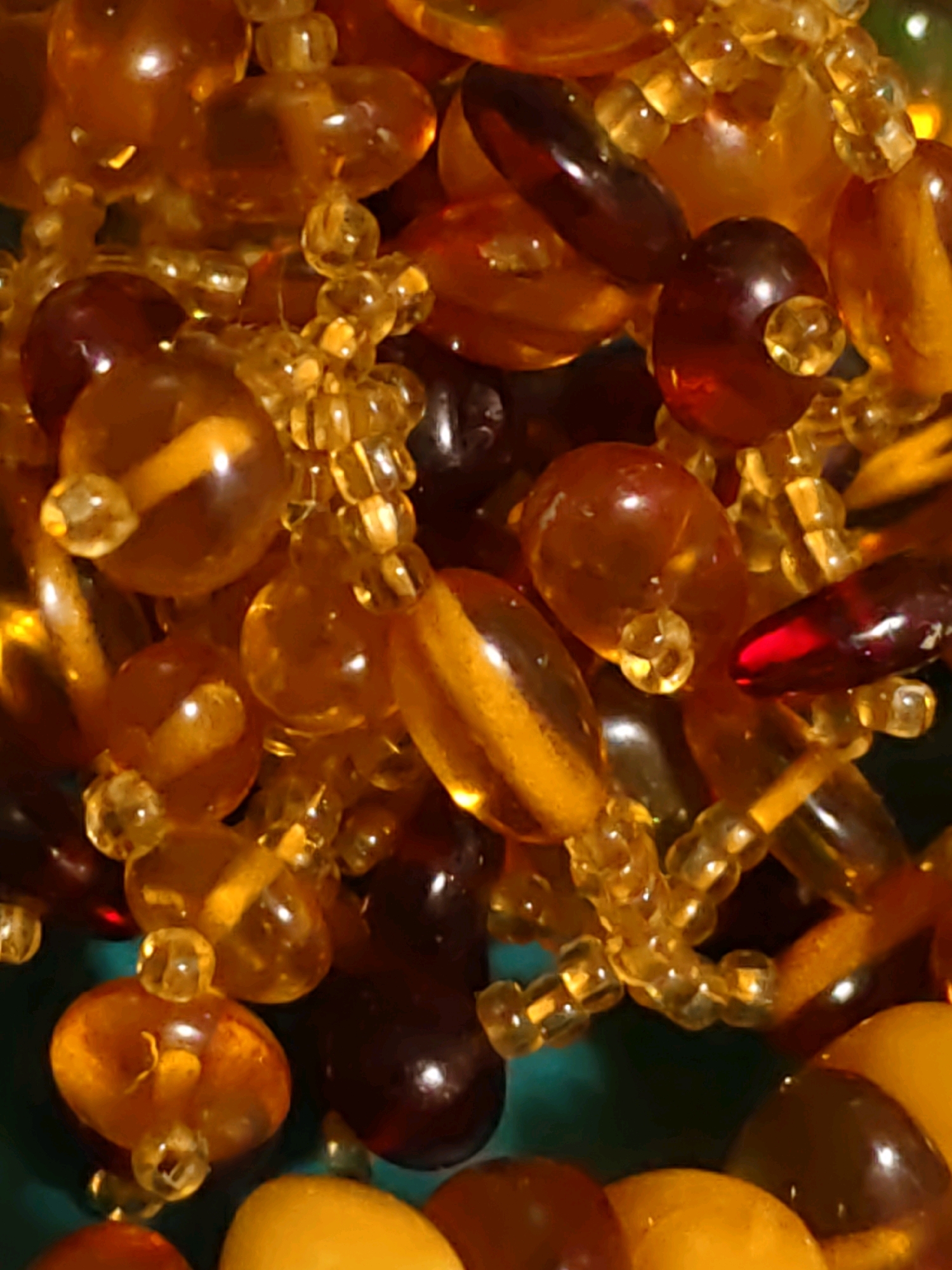 The image is a close-up, overhead shot of a collection of amber beads. The beads are various sizes and shapes, including round, oval, and irregular. They range in color from light yellow to deep brown, with some appearing translucent and others more opaque. Many of the beads are smooth and polished, reflecting light. Some smaller, clear beads are strung between the larger amber pieces, creating a necklace-like effect. The background is a dark, out-of-focus teal color. There are no people or specific landmarks visible that would suggest a location in Rostov-on-Don, Russia. The image appears to be an artistic or decorative arrangement of jewelry.