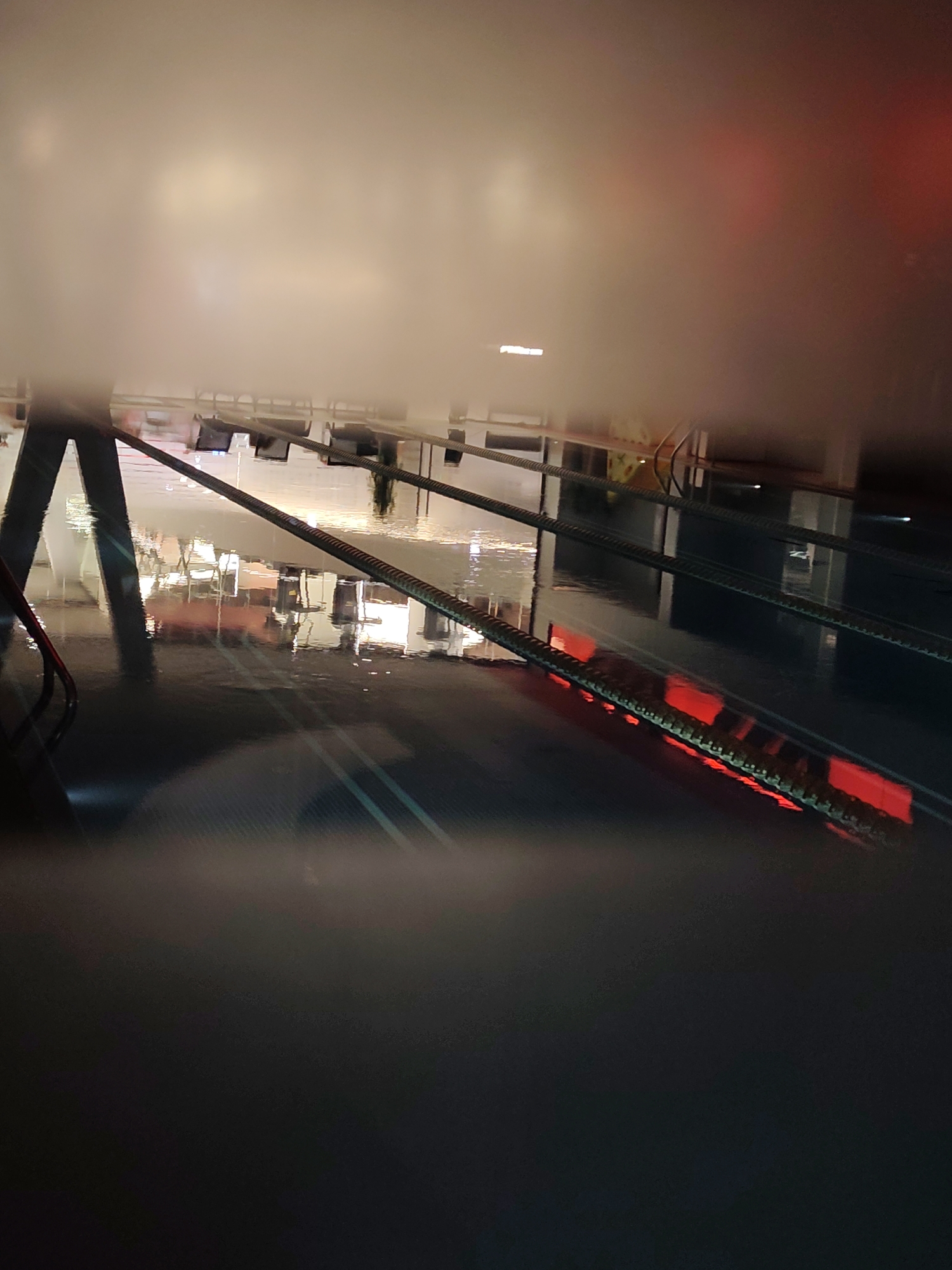 The image shows an indoor swimming pool in Moscow, Russia at night. The pool lanes are separated by ropes with red floats. Reflections of chairs and tables are visible in the water. The scene appears hazy, possibly due to humidity.