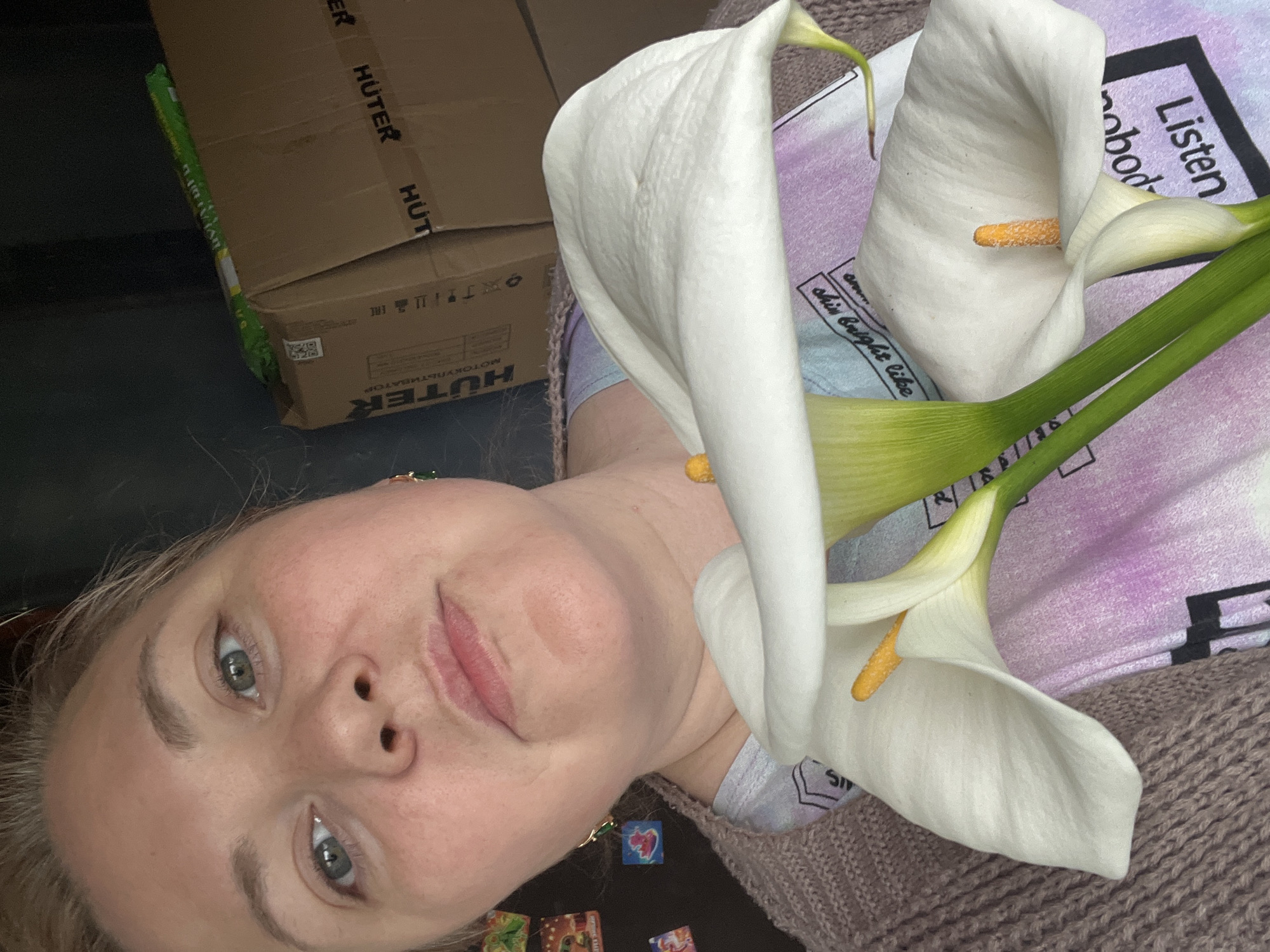 A person is holding three white calla lilies in front of them. The person has fair skin, light hair, and gray eyes. The background includes a cardboard box with the word "HUTER" printed on it, a patterned shirt, and a brown knitted sweater. There is no indication of Vesyoloye, Russia in the image.