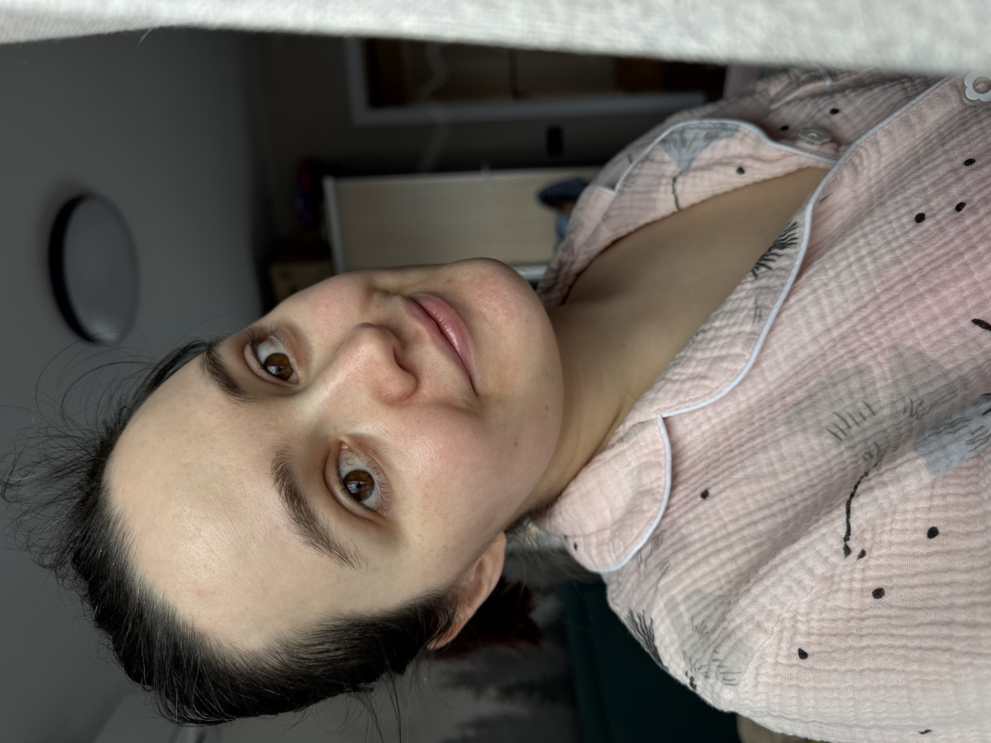 A selfie taken indoors, showing a woman with fair skin, dark hair pulled back from her face, and brown eyes, wearing a light pink pajama top with a white trim and a small black pattern. She is looking slightly upwards at the camera with a neutral expression. Behind her is a blurry background with a light-colored wall, a circular object mounted on the wall, and a glimpse of furniture. The image does not provide sufficient information to determine the specific location in Izhevsk, Russia.