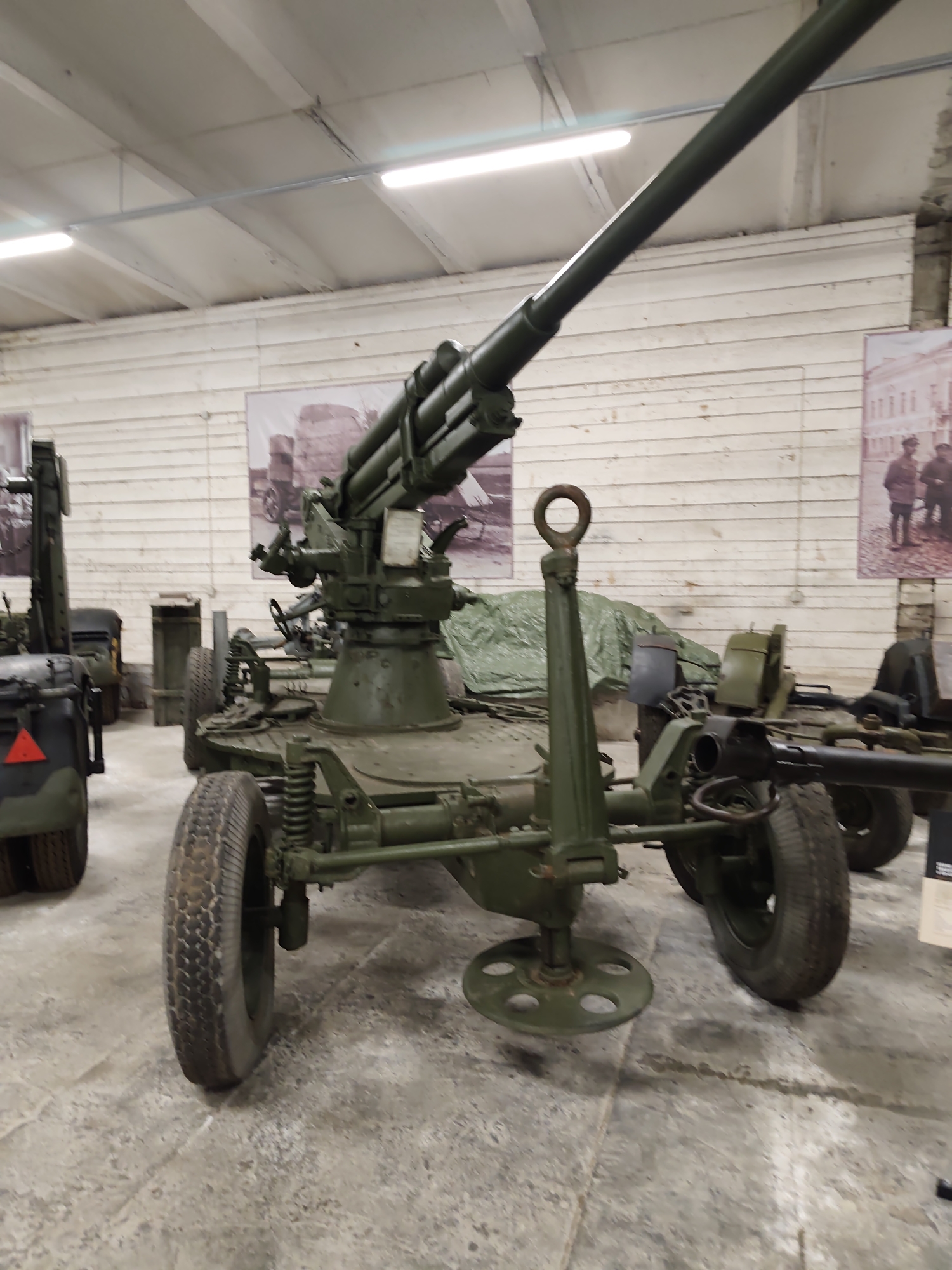 The image depicts an outdoor display of military artillery at the Estonian War Museum – General Laidoner Museum in Viimsi, Estonia. The primary subject is a green, wheeled anti-aircraft gun positioned centrally in the frame. The gun features a long barrel, a rotating turret with various control mechanisms, and a stabilizing leg with a circular base that rests on the ground. The gun is mounted on a chassis with two large tires visible in the foreground and rear. In the background, a white wooden wall serves as a backdrop, adorned with two large black and white photographs. The left photograph shows a military vehicle, while the right depicts several figures in uniform. To the left of the main artillery piece, another military vehicle, partially visible, has a red triangular marker. The ground is a concrete surface, showing some wear and tear. The museum environment is suggested by the indoor setting with industrial lighting visible on the ceiling.