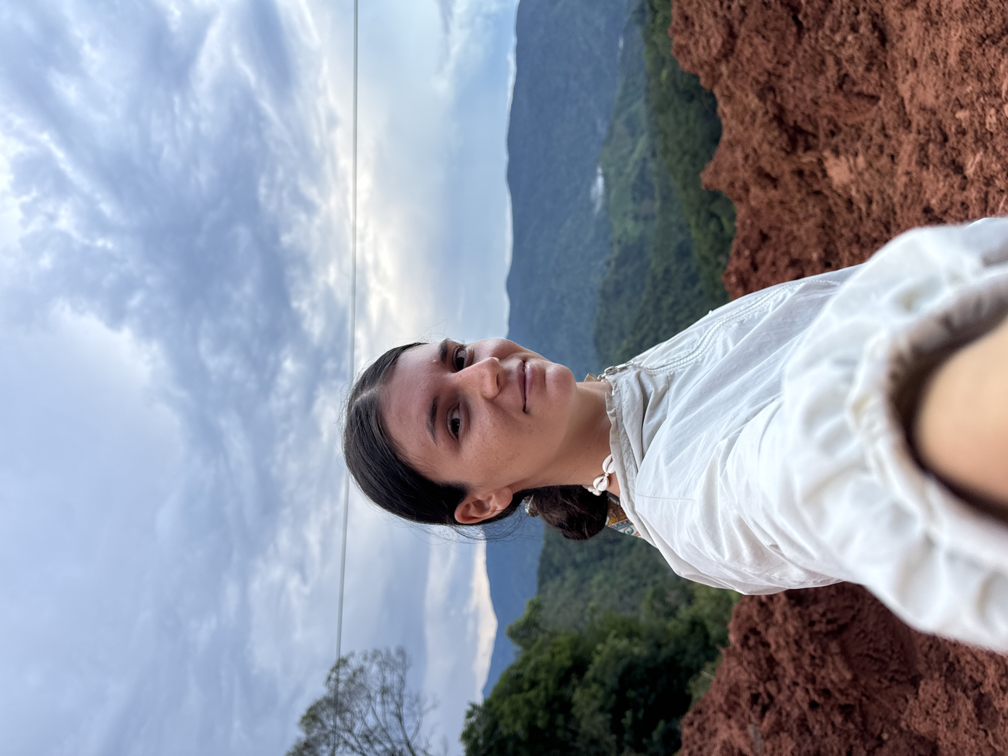 A young woman with dark hair pulled back is taking a selfie in a mountainous, rural landscape. She is wearing a light-colored jacket and a shell necklace. Behind her, rolling green hills and mountains stretch into the distance under a cloudy sky. In the foreground, mounds of reddish-brown soil are visible. The scene appears to be located in or near the city of Nao Sẻ, Vietnam, indicated by the lush, tropical mountain vegetation and the type of soil.