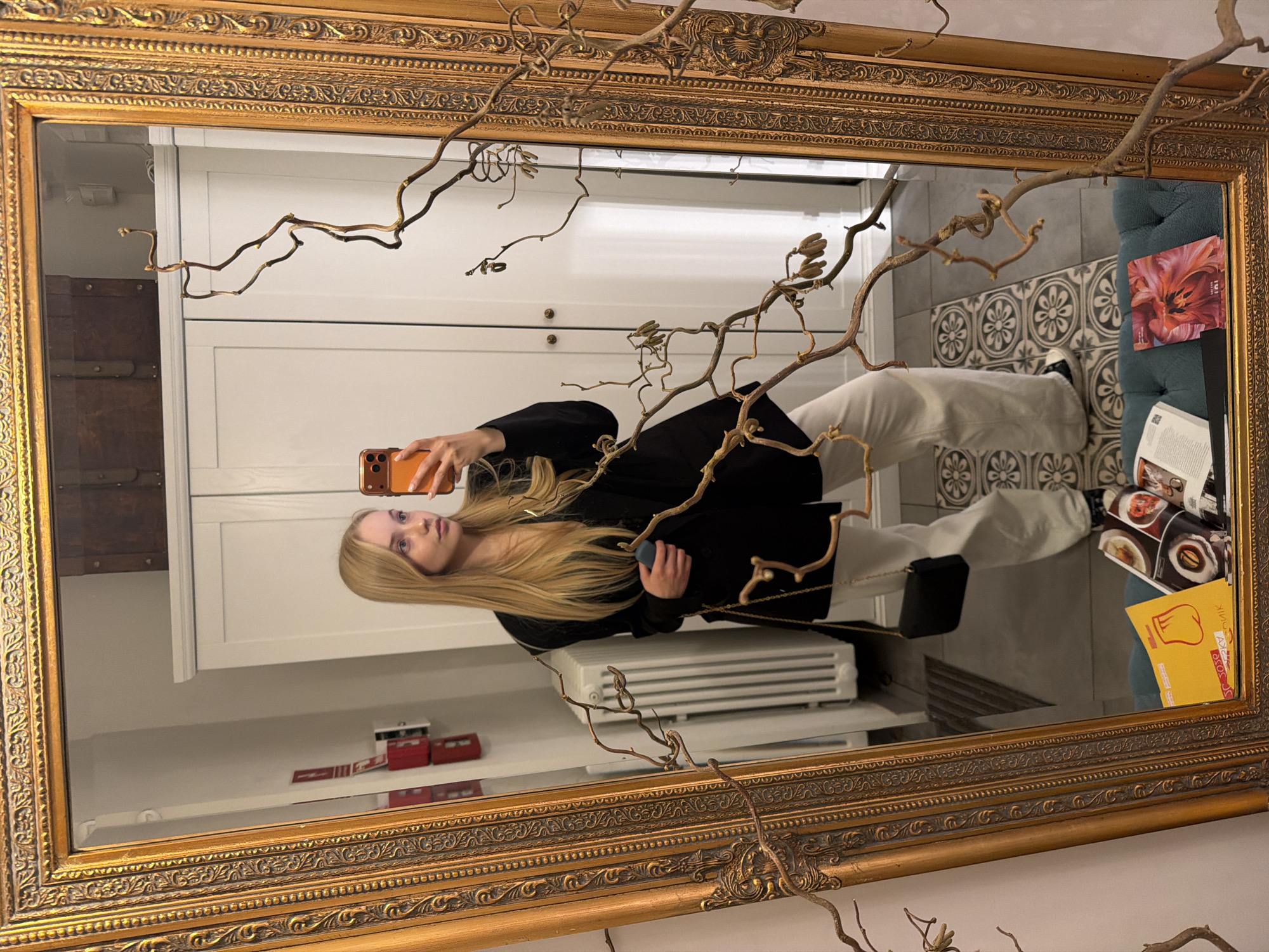 The image depicts a young woman taking a selfie in a large, ornate gold-framed mirror. The mirror reflects a hallway or entryway with white doors, a radiator, and a shelf with small objects. Behind the woman, there is a patterned tile floor and a portion of a blue upholstered seat with books and papers on it. The mirror frame is partially obscured by dried branches. The location appears to be an interior setting within the city of Unknown, Unknown.