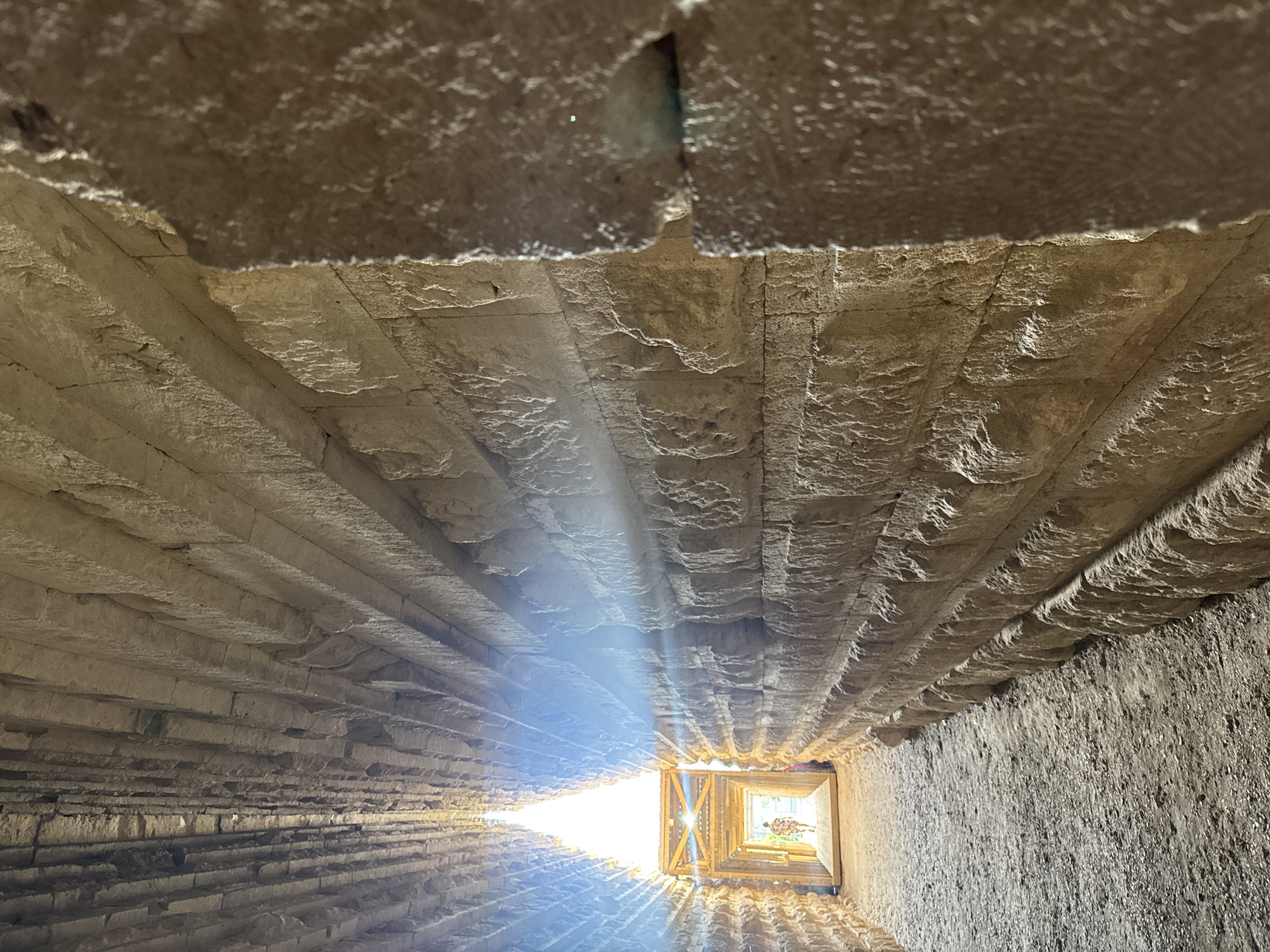 The image depicts an interior view of a long, narrow corridor constructed with large, rough-hewn stone blocks. The corridor walls are textured and uneven, with visible striations and cracks in the stonework. Sunlight streams in from a bright opening at the far end, creating a strong backlight effect and illuminating dust motes in the air. The ground appears to be covered in gravel or dirt. At the exit of the corridor, a wooden structure with visible beams is present, and a person is walking away from the camera towards the light. The scene suggests an ancient structure or archaeological site, potentially a tomb or passageway, located in Kerch, Ukraine.
