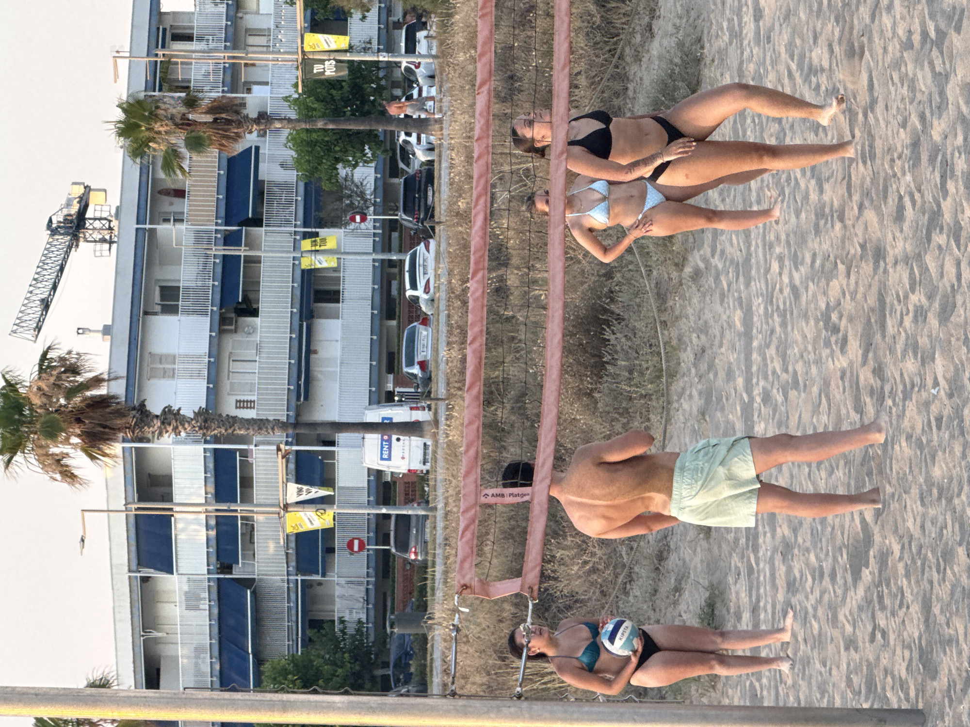 This is a vertical, wide-angle photograph taken at beach level, showing a scene with people playing beach volleyball. The image is oriented such that the subjects and background elements are upside down relative to a standard upright orientation.
In the foreground, the beach volleyball net divides the frame. On one side of the net, a woman in a turquoise bikini is holding a volleyball, appearing to be either preparing to serve or receive. On the other side of the net, a shirtless man is visible, wearing light green shorts. Further in the background, two women in bikinis are playing volleyball. One wears a black bikini and the other a light blue bikini.
Behind the beach volleyball court, a dry, sandy area with sparse vegetation is visible, leading to a tall, multi-story building with balconies. A construction crane is positioned to the left of the building. Palm trees are scattered throughout the scene.
The lighting suggests it is daytime, likely late afternoon or early evening, given the warm tones and soft shadows on the sand. The sky appears bright and clear, indicating fair weather.
Visible text includes "RENT O" on a white van in the background, and smaller yellow signs on the building. There are also markings on the volleyball, including "KIPSTA". The volleyball net itself has a red strap with text that appears to read "AMB Platges".
The overall atmosphere of the image is relaxed and active, capturing a moment of recreational sports on a beach in a coastal urban environment, possibly Castelldefels, Spain, as indicated by the location context.