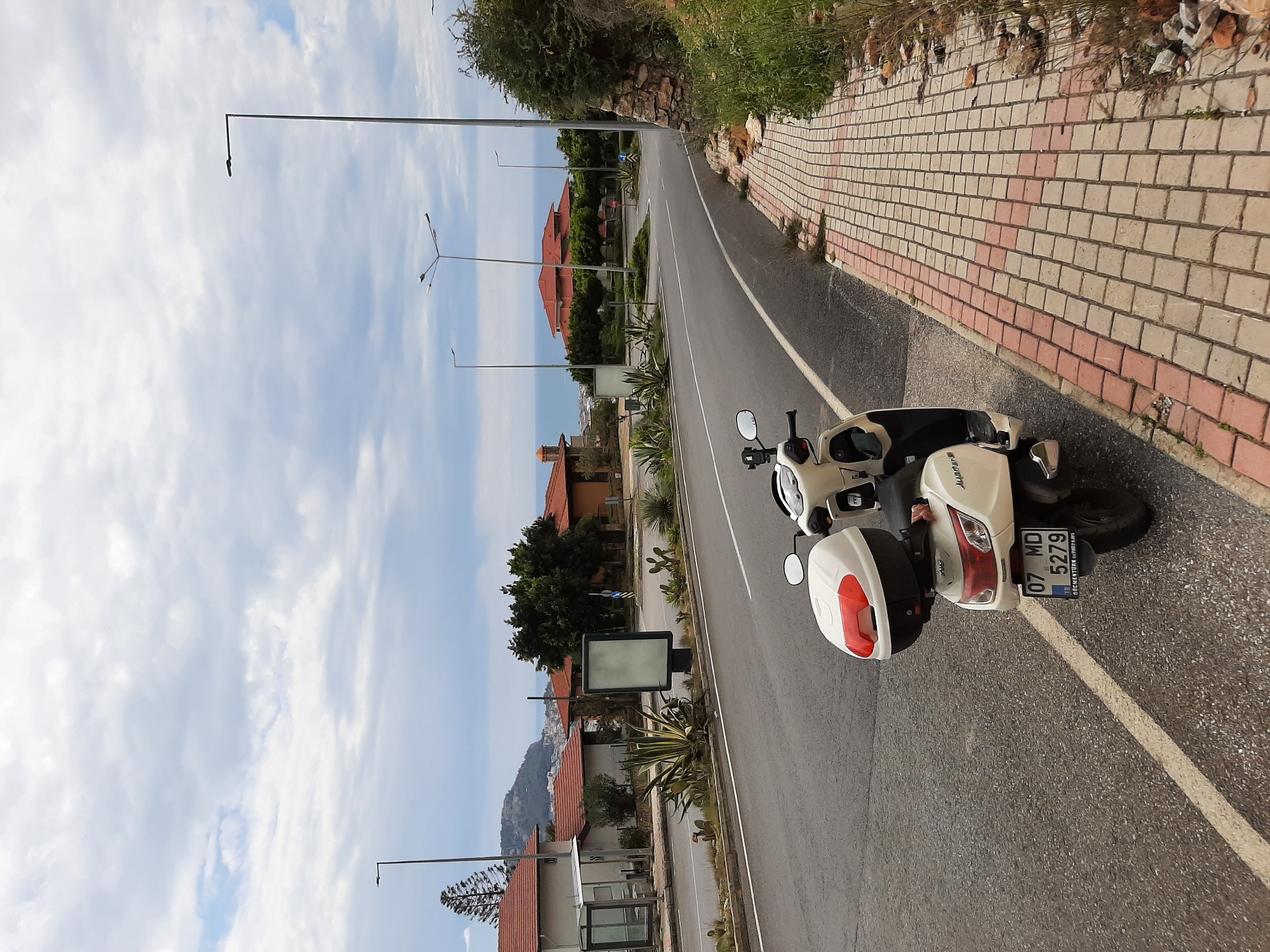 A moped is parked on the side of a road in Alanya, Turkey. The moped is white with a red and black storage box on the back. The license plate number is 07 MD 5279. The road is asphalt with a white line marking the edge. A brick sidewalk borders the road. Buildings and trees line the other side of the road. There are streetlights and a billboard nearby. In the distance, a mountain and the sky are visible.