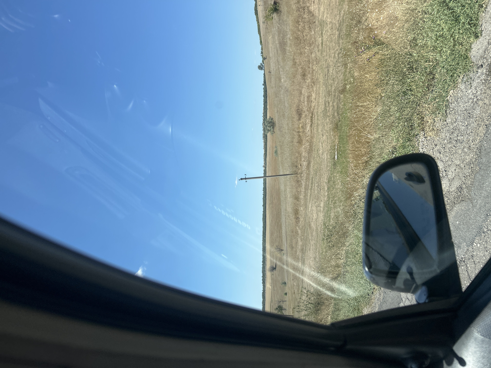 The image depicts a dry, grassy landscape under a clear blue sky, as viewed from the interior of a car. The foreground shows the interior of the car, including the car's side mirror reflecting the sky and a portion of the car. The side mirror is black and mounted on the car's frame. The exterior view consists of a vast expanse of dry, yellowed grass and sparse vegetation, indicating a dry season. A single wooden utility pole stands in the middle ground, and a line of trees is visible in the far distance along the horizon. The terrain appears to be gently rolling hills. There are no people visible in the scene, and the overall impression is one of a rural, possibly steppe-like environment. The image was taken in the city of Cioc-Maidan, Moldova.