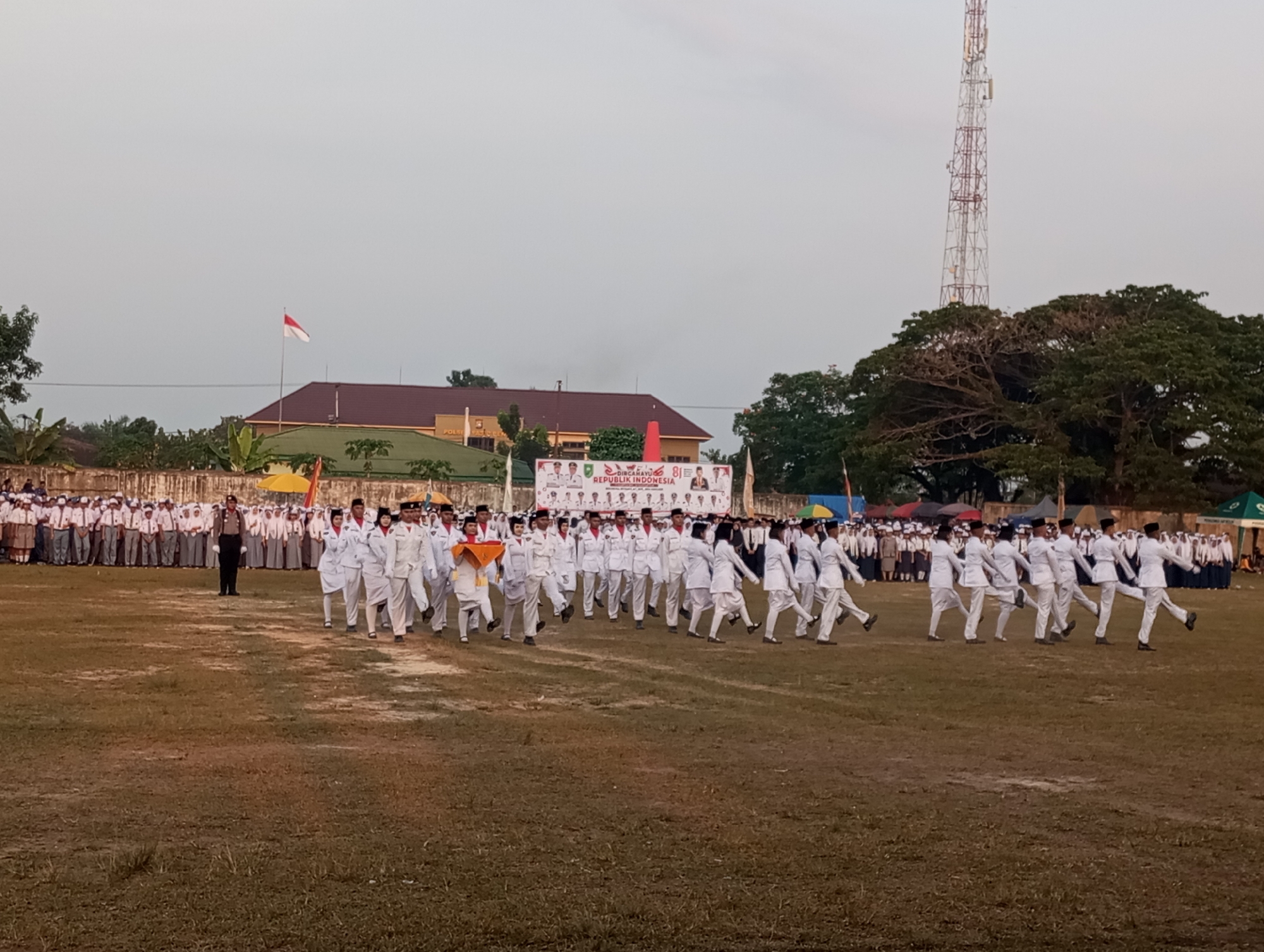 The image depicts an outdoor event, likely a parade or ceremony, taking place in Candirejo, Indonesia. In the foreground, a group of individuals in white uniforms with red sashes are marching in formation across a grassy field. Their posture suggests they are performing a synchronized march. Behind them, a larger group of people in similar white uniforms and brown skirts or pants are standing in rows, observing the marchers. A lone figure in a dark uniform, possibly a police officer, stands facing the marching group. In the background, a building with a red roof and banners displaying text, including "REPUBLIK INDONESIA," can be seen. A tall telecommunications tower is visible to the right, and a large tree with dense foliage stands behind it. The sky is overcast, indicating it might be late afternoon or early morning. The overall scene suggests a patriotic or ceremonial event.