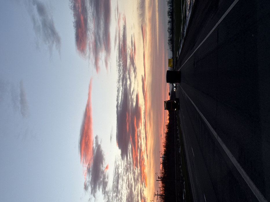 The image, oriented vertically but depicting a horizontal landscape, captures a serene yet dramatic sunset or sunrise scene from the perspective of someone traveling on a multi-lane highway.
The left half of the image is dominated by a magnificent sky, transitioning from a soft, pale blue at the top to a vibrant tapestry of orange, pink, and yellow closer to the horizon. Streaky, elongated clouds are painted with intense hues of red and orange, particularly in the lower and middle portions of the sky, contrasting with pockets of lighter white and gray clouds. The overall effect suggests the magical hour of twilight.
The right half of the image features a dark asphalt highway, seemingly stretching into the distance. White lane markings clearly delineate the travel lanes and a shoulder on the right side. The road surface appears dark, possibly due to low light conditions or a slight dampness, reflecting minimal light.
Several vehicles are visible on the highway. In the mid-ground, a dark silhouette of a large vehicle, possibly a truck or a semi-trailer, is prominent against the brighter horizon. Further down the road, a second, lighter-colored vehicle, which appears to be a semi-truck, is clearly distinguishable with its yellow or amber headlights glowing softly, illuminating the path ahead. In the far distance, faint pinpricks of light indicate other vehicles.
Along the horizon, separating the colorful sky from the highway, is a dark, silhouetted landscape. This appears to be a treeline or dense vegetation bordering the road. On the left side of the highway, a series of utility poles or similar structures are visible in silhouette against the vibrant sky.
The time of day is clearly either sunrise or sunset, given the striking colors in the sky and the low ambient light, which necessitates the use of vehicle headlights. The atmosphere is calm and contemplative, emphasizing the beauty of nature even amidst infrastructure like a highway. No discernible text is visible in the image.