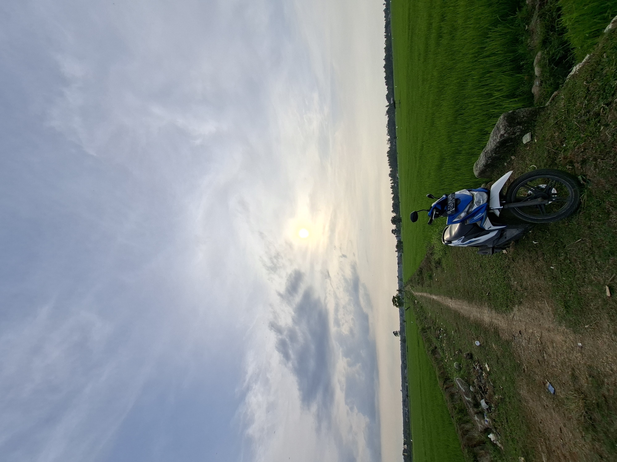 A blue and white Honda scooter is parked on a dirt path next to a vibrant green rice field. The path runs between the field and a ditch. The sky is a soft blue with scattered white and grey clouds, and the sun is visible, peeking through the clouds. The scene appears to be in a rural area, likely Kayeepanyang, Indonesia, given the presence of rice paddies and the scooter, a common mode of transportation in the region. The atmosphere is calm and serene, with the lush green field stretching out into the distance.