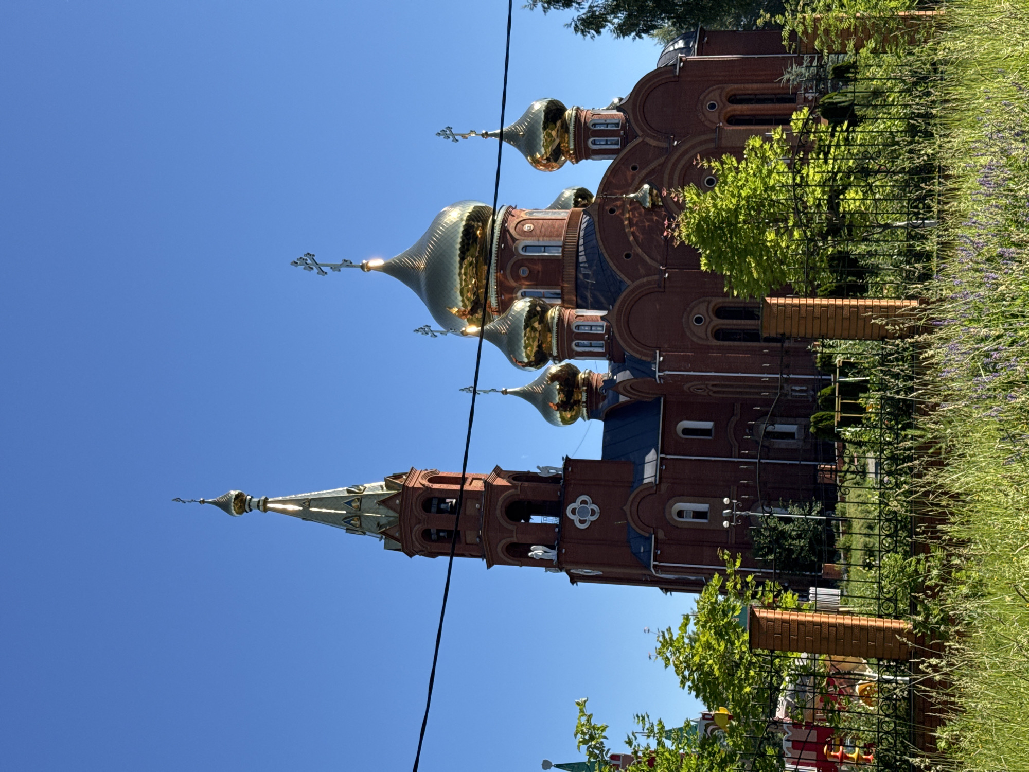 The image shows an Orthodox church, likely the Church of the Archangel Michael, in Chaikovsky, Russia. The church is constructed of red brick with multiple golden domes. A tall bell tower with a spire is visible on the left side of the building. The sky is clear and blue. In the foreground, there is a black metal fence, with tall grass and some flowering plants growing behind it. A power line runs horizontally across the upper portion of the image, obscuring part of the sky and the church. To the left of the main church building, a portion of another church with a green spire can be seen.