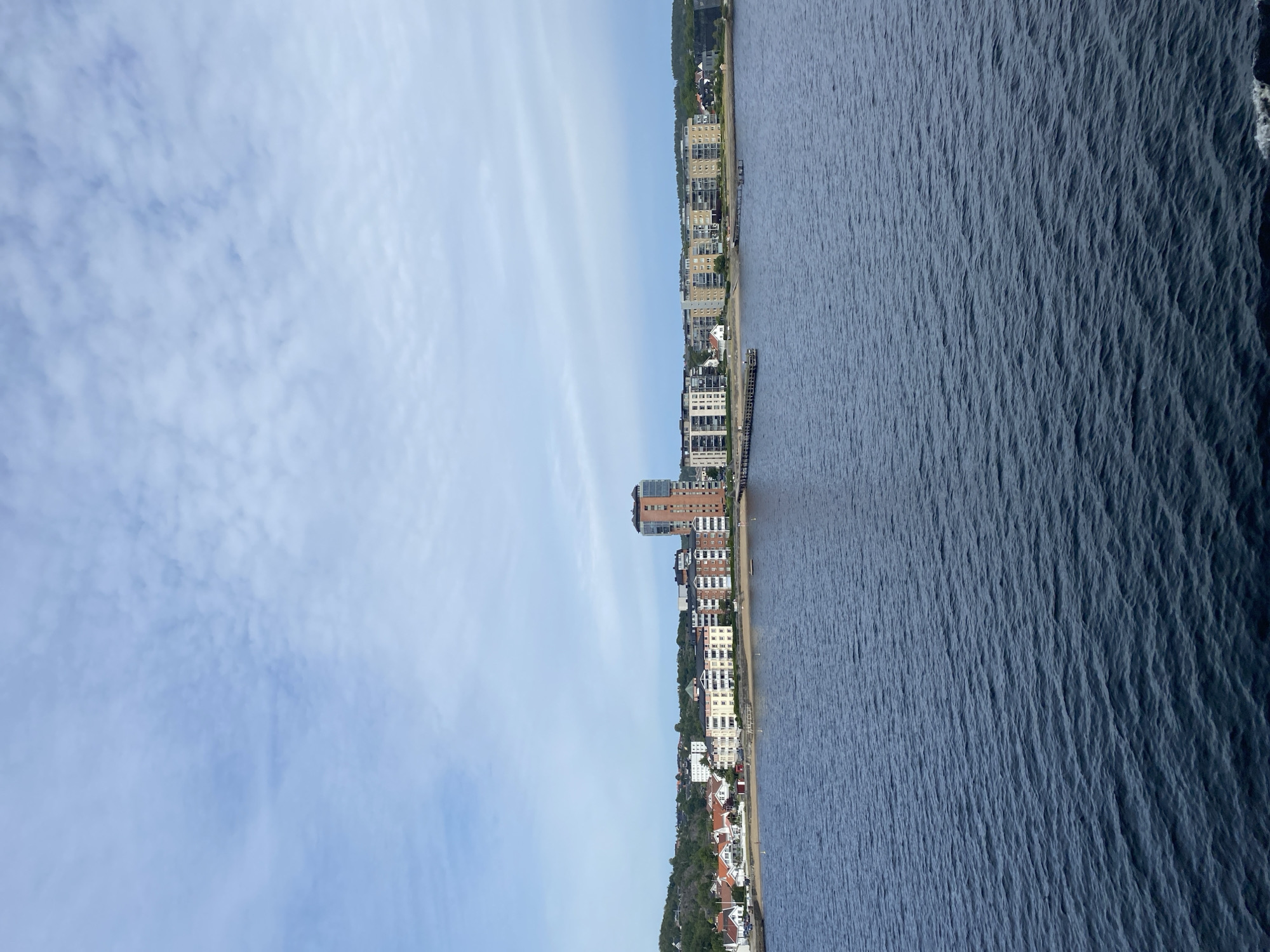 The media file depicts a scenic view of the coast in Larvik, Norway. The image is oriented vertically, with the sky occupying the left half and the sea and shoreline on the right. Wispy white clouds are scattered across a pale blue sky, suggesting it is daytime. The sea is a deep blue, with a textured surface indicative of gentle waves.
Along the shoreline, a series of buildings is visible. These range from modern, multi-story apartment complexes to smaller houses with red-tiled roofs, nestled amongst lush green trees that cover the rolling hills. A pier or jetty extends into the water from the shore, serving as a docking point. The architecture suggests a developed urban or suburban area situated next to the water. The overall atmosphere is serene and picturesque, capturing a typical coastal day in a Norwegian town. No people are prominently visible, and the focus is on the natural landscape and the built environment.