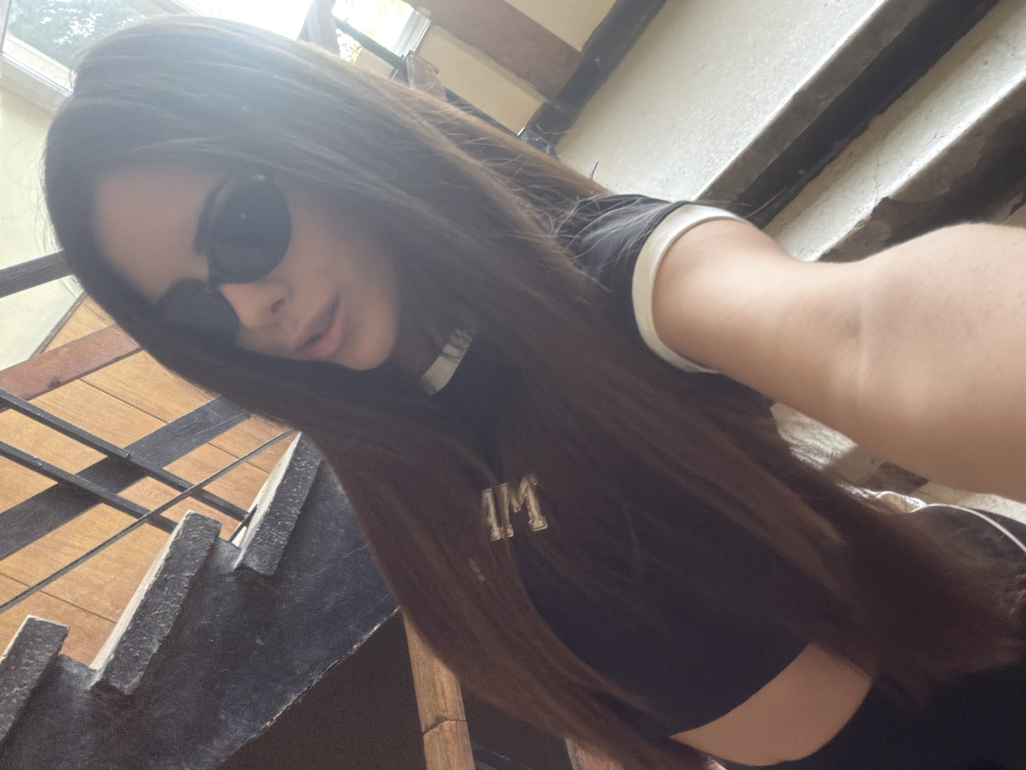 A woman is taking a selfie while standing on wooden stairs with metal railings. She is wearing sunglasses and a black crop top with white trim, which has the letters "WM" on the front. The woman has long, dark hair. In the background, there are concrete stairs. The location is implied to be Sofia, Bulgaria.