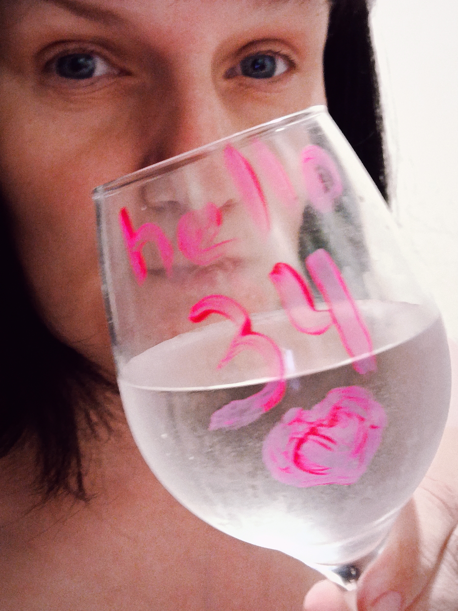 A close-up, eye-level shot features a person, likely female, holding a clear stemware glass in front of their face. The person's blue eyes are visible above the rim of the glass, looking directly at the camera, while dark hair frames the right side of the face. The background is a plain, light surface, suggesting an indoor setting.
The glass contains a clear liquid, possibly water, filling less than half its volume. Bright pink writing is prominently displayed on the glass, spelling "hello 34" and including a small, stylized heart or swirl design towards the bottom. The writing appears to be applied with paint or marker. The image has a slightly grainy texture.
The scene depicts a personal moment, possibly a celebration or acknowledgement of a birthday or milestone (implied by "34"). The person's expression is neutral to calm. The location context provided for this media is Фоки, Russia.