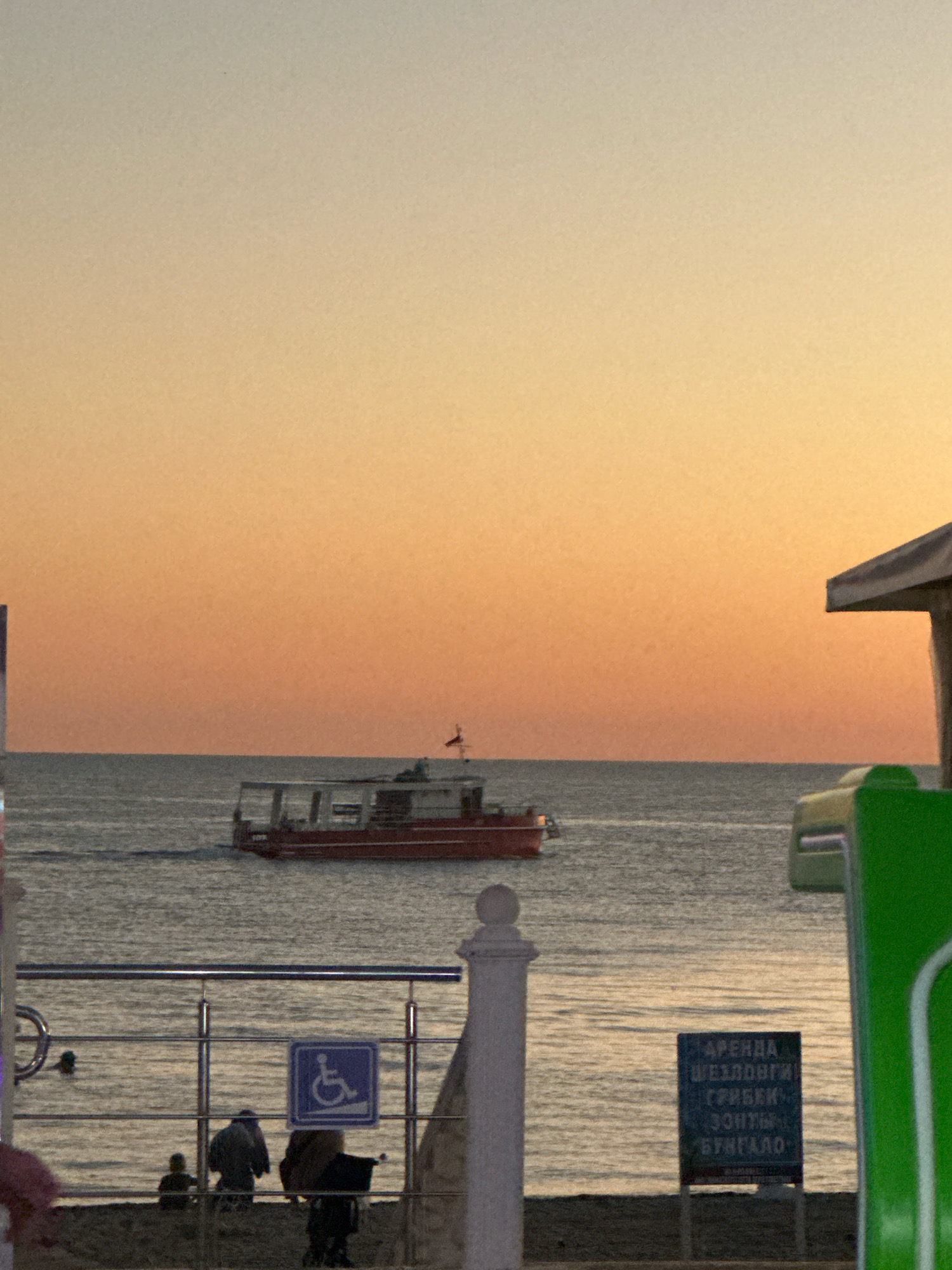 The image depicts a sunset scene at a beach in Lermontovo, Russia. The sky is a gradient of orange and yellow. A red boat with a white roof is sailing on the calm sea. In the foreground, there is a beach area with a metal railing. A blue sign with a wheelchair symbol is visible. There are several figures on the beach, some appearing to be sitting. A dark sign with Russian text is also present. To the right, part of a green structure and a light-colored roof are visible, suggesting a building or an amenity at the beach.