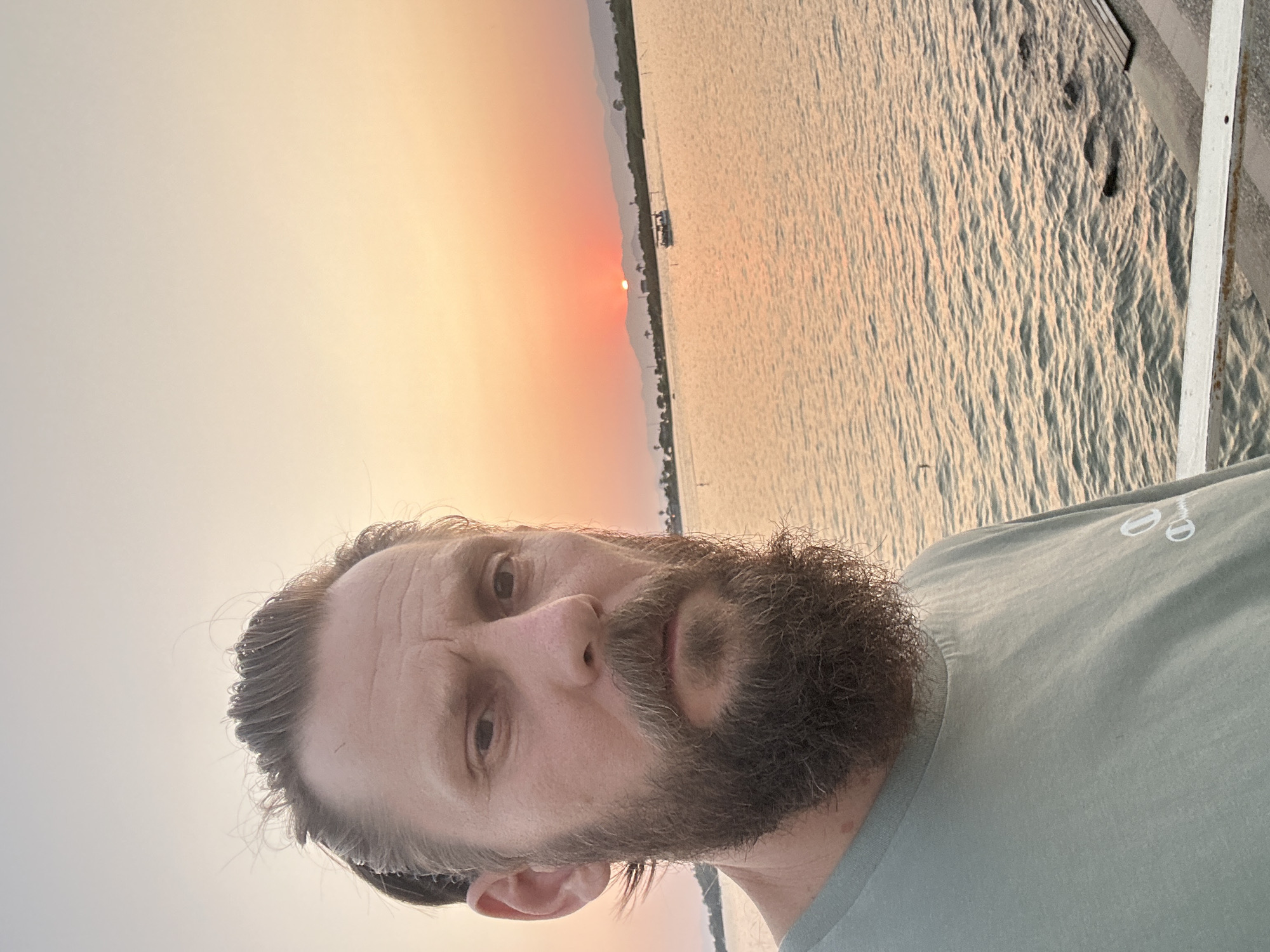 A selfie taken at sunset shows a Caucasian man with light brown hair pulled back and a full beard. He is wearing a light green t-shirt. The man is positioned in the foreground, slightly to the left of the frame. Behind him, a body of water reflects the warm hues of the setting sun, with a silhouette of a distant shoreline dotted with buildings and mountains. A small boat with people on board is visible on the water. The overall atmosphere is tranquil, with a gentle breeze rustling the man's hair. The location appears to be a waterfront area, possibly a pier or promenade, in the city of Fethiye, Turkey, given the characteristic sunset over the Aegean Sea.
