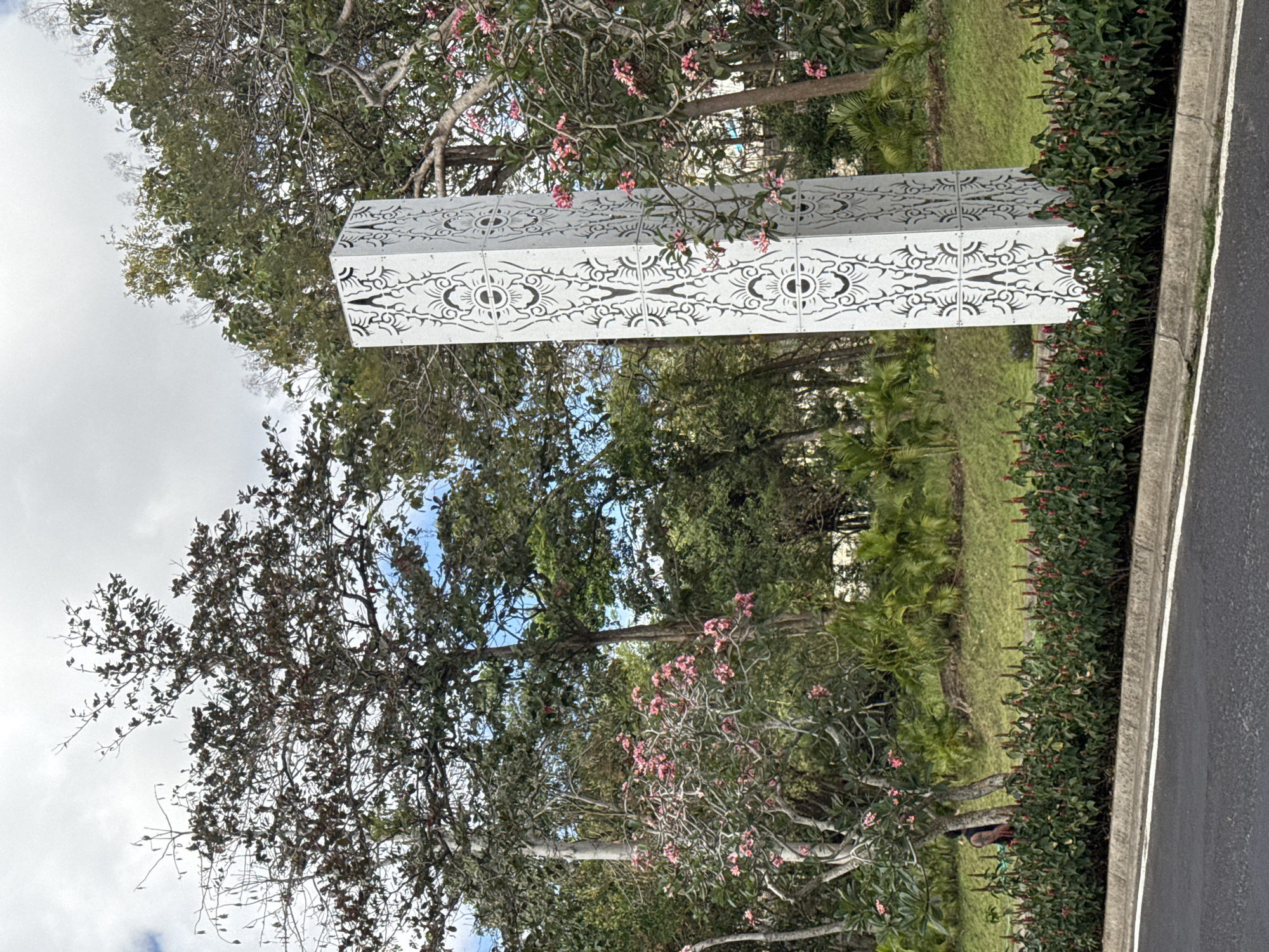 A tall, white, rectangular sculpture with intricate black geometric patterns stands on a grassy median next to a road. The sculpture is illuminated by daylight, and its designs appear to be carved or cut out of the material. Behind the sculpture, a variety of trees and lush green vegetation can be seen, some with pink flowers. A person with dark hair and tattoos on their arms is partially visible in the background, standing behind a row of small, red-flowered bushes. The sky above is partly cloudy. This scene is located in Kuta Selatan, Indonesia.