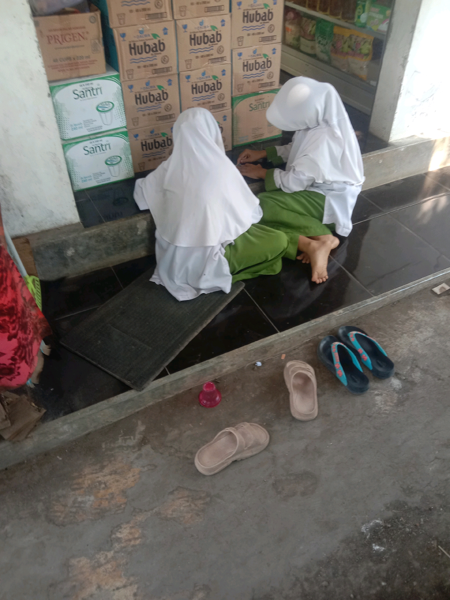 In Ngempit, Indonesia, two young individuals wearing white hijabs and green skirts are seated on a tiled floor. One is positioned behind the other, facing away from the camera, with their head bowed. In front of them, a collection of stacked cardboard boxes are visible, some bearing the brand name "Hubab" and others "Santri." To the left, a portion of a red and pink patterned fabric is visible, likely clothing. In the foreground, several pairs of sandals are scattered on the concrete ground: two pairs of flip-flops, one black with blue and red accents, and another pair of tan slip-on sandals. A small pink object, possibly a toy, is also on the ground near the tiled area. The environment appears to be an outdoor space, possibly a storefront or a porch, with sunlight casting shadows.