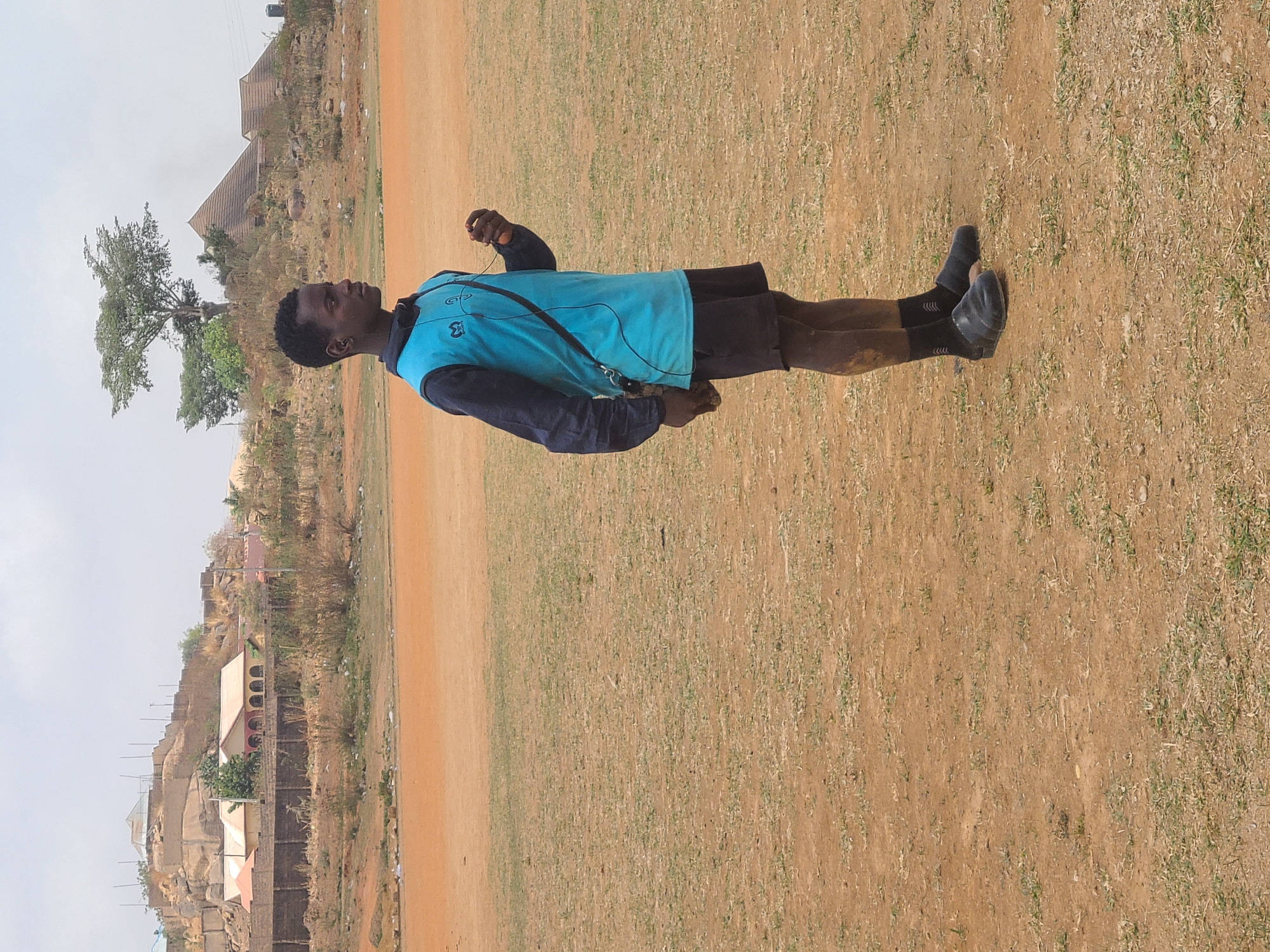 A medium shot captures a young Black man standing in a field in Barikin Jirga Summa, Nigeria. He faces left, dressed in a light blue vest over a dark blue shirt and dark shorts. He wears dark socks and shoes. A black cord, likely from headphones, is draped across his chest.
The field consists of brown dirt and sparse green grass. In the background, structures sit on rocky terrain, a tree with a wide canopy stands to the left of the man. The sky is overcast.