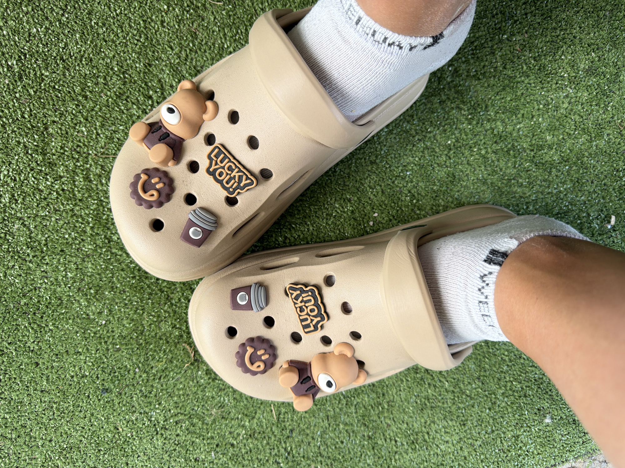 A pair of tan-colored Crocs, adorned with cartoonish charms of a bear, a flower, a spray can, and the words "LUCKY YOU!", are worn by an individual whose feet, clad in white socks, rest on a patch of artificial turf. The scene is outdoors, with the artificial turf providing a green, textured ground cover. The lighting suggests it is daytime. No specific location in the city of Rusokastro, Bulgaria, can be identified from the image.