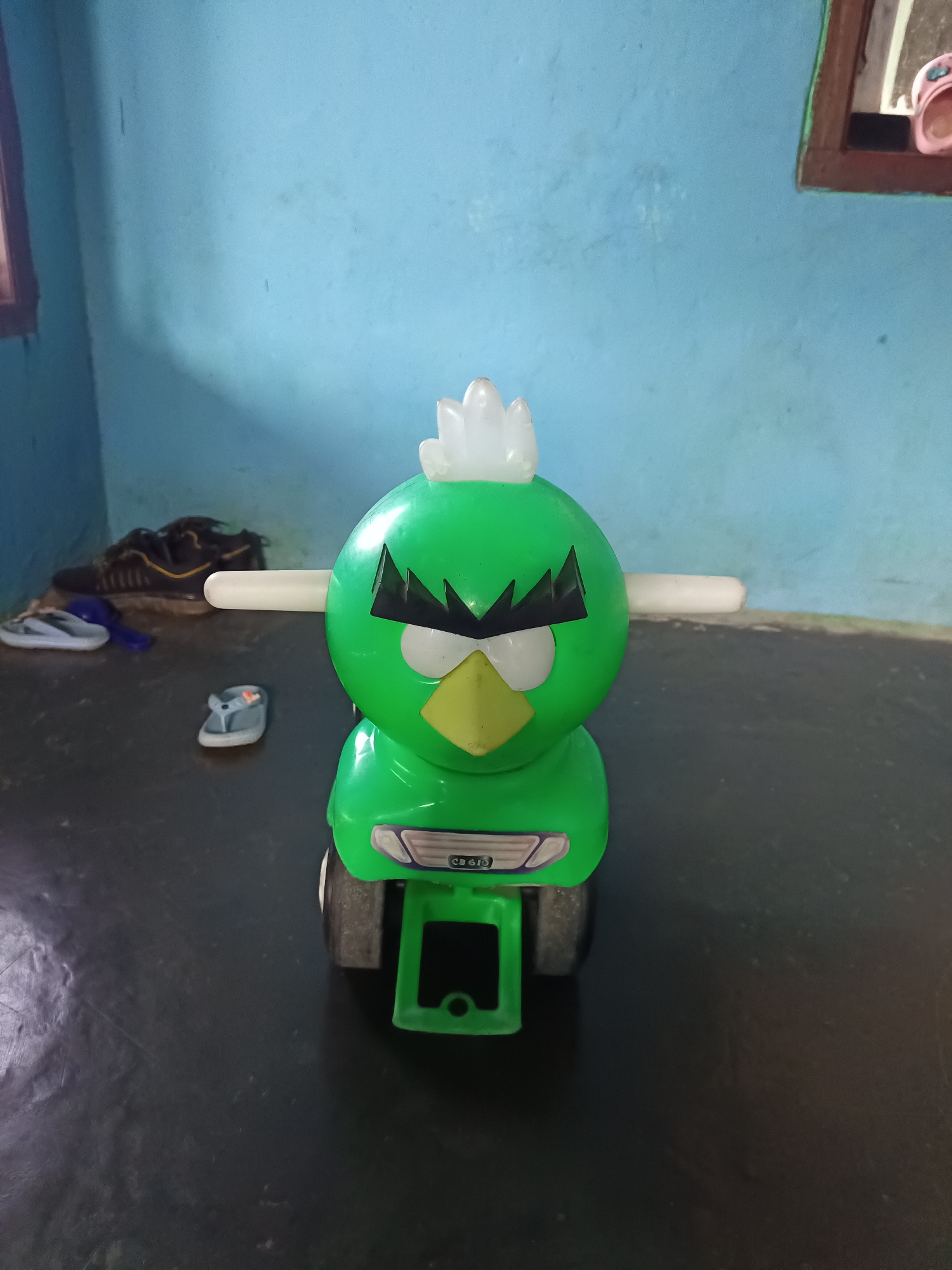 A close-up, eye-level, frontal shot focuses on a bright green plastic toy. The toy resembles a cartoon bird with a white feathered crest, black angry eyebrows, and a yellow beak. It has two white cylindrical rods protruding from its sides. The toy is positioned on a dark, textured floor. In the background, a light blue wall is visible, with a wooden window frame in the upper right corner. Part of a pink slipper can be seen within the window frame. To the left of the toy, a pair of dark athletic shoes and a pair of gray flip-flops are on the floor. The overall scene suggests an indoor environment, possibly a child's room, within a residential setting in Terbanggi Marga, Indonesia.