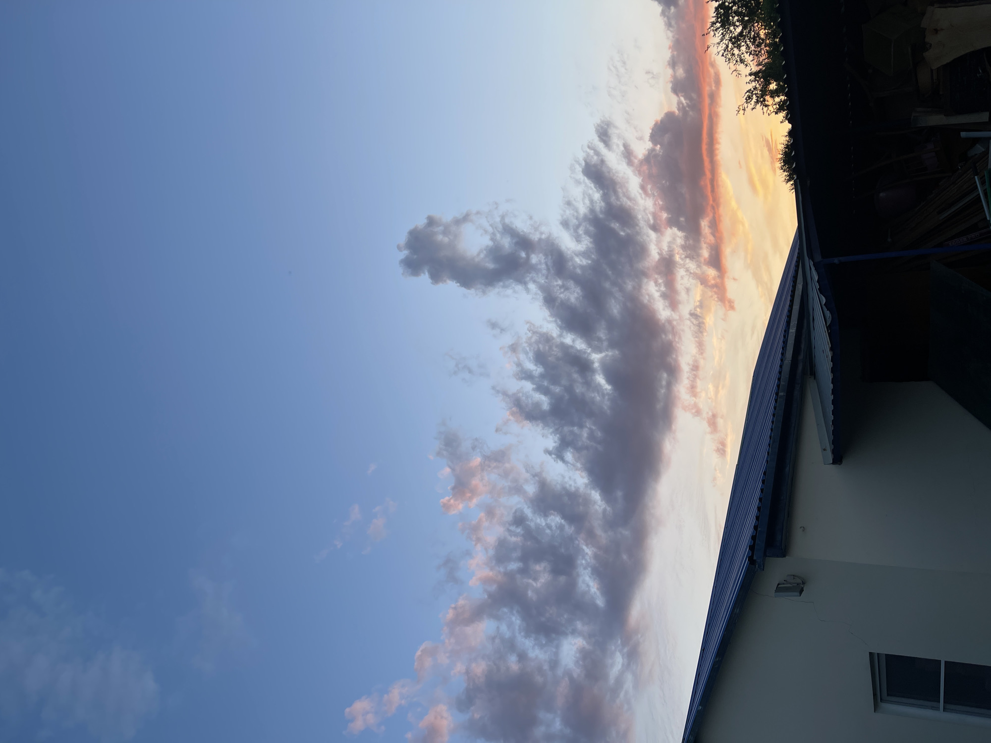 The image depicts a twilight sky over a residential area. The sky is a gradient of blue, transitioning to pink and orange hues near the horizon, with scattered clouds. In the foreground, the roof of a building is visible, along with a covered outdoor storage area. The building appears to be a light-colored, single-story structure with a window. The storage area contains various objects, including what appear to be lumber and containers. The scene is located in the city of Donskoye, Russia.