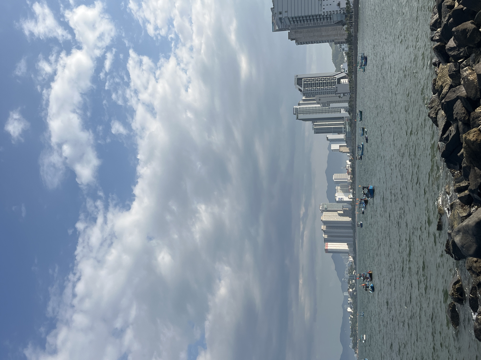 The image captures a coastal urban landscape in Bac Nha Trang Ward, Vietnam, under a partially cloudy sky. The upper sky features patches of clear blue with scattered white cumulus clouds, while the mid-sky is dominated by a more uniform layer of grey clouds. The foreground consists of large, dark grey to black boulders forming a rocky shoreline, with small waves breaking against them. The dark green-grey sea occupies the midground, dotted with approximately 12 to 15 small, traditional Vietnamese fishing boats, primarily colored blue, green, and red/yellow, which are either anchored or adrift. In the background, a dense urban skyline of modern multi-story buildings, predominantly white and grey high-rise residential or hotel towers, extends along the coast. Hazy, undulating mountains rise directly behind the city buildings. No individual people are distinctly visible.