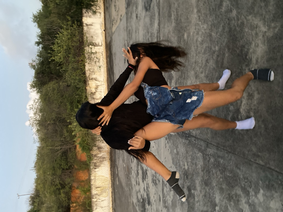 Two figures are tangled in a potential embrace. One figure, clad in black long sleeves and head covered, seems to be holding the other. The second figure wears a dark tank top and a short, denim skirt. One figure wears white socks, while the other sports black socks. They are on a grey surface, with greenery visible in the background and sky to the side.