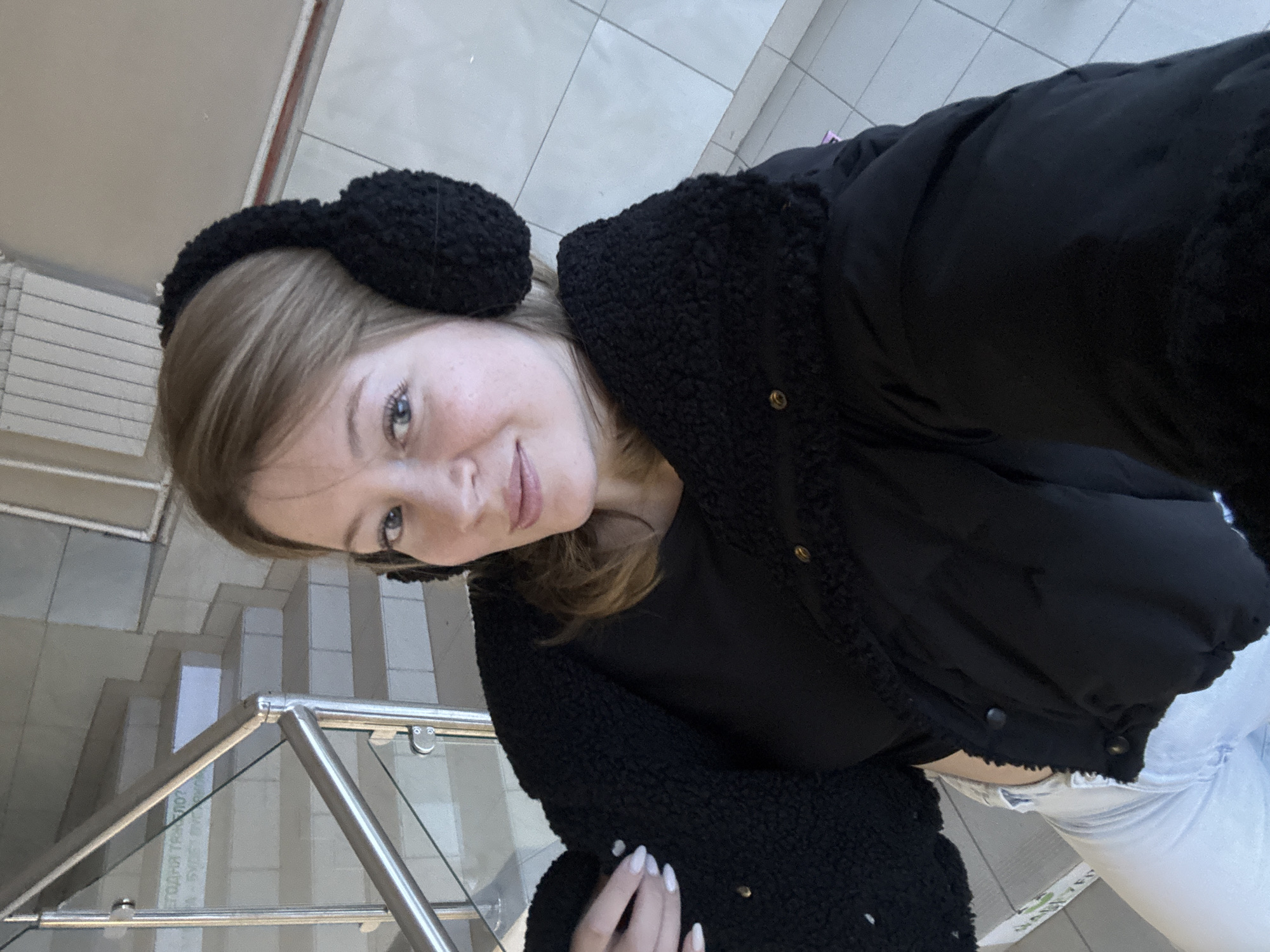 A young woman is taking a selfie indoors. She is wearing black ear muffs and a black puffy jacket with a black shirt underneath and light-colored jeans. Her nails are painted white. The background appears to be a stairwell with a metal and glass railing. The location is Chaikovsky, Russia, as evidenced by the Cyrillic text visible in the background.