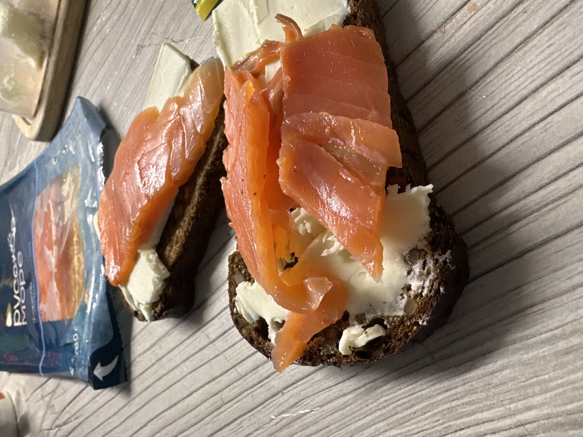 The image shows two open-faced sandwiches on a light gray, wood-grain patterned surface, likely a table. Each sandwich consists of a slice of dark rye bread spread with a creamy white substance, possibly cream cheese. Topped with several pieces of sliced, orange-colored smoked salmon. An opened package of smoked salmon is visible to the left of one sandwich. The text on the package is in Cyrillic script. The location is likely in Chaikovsky, Russia, as the text on the salmon package "русское море" is a Russian brand.