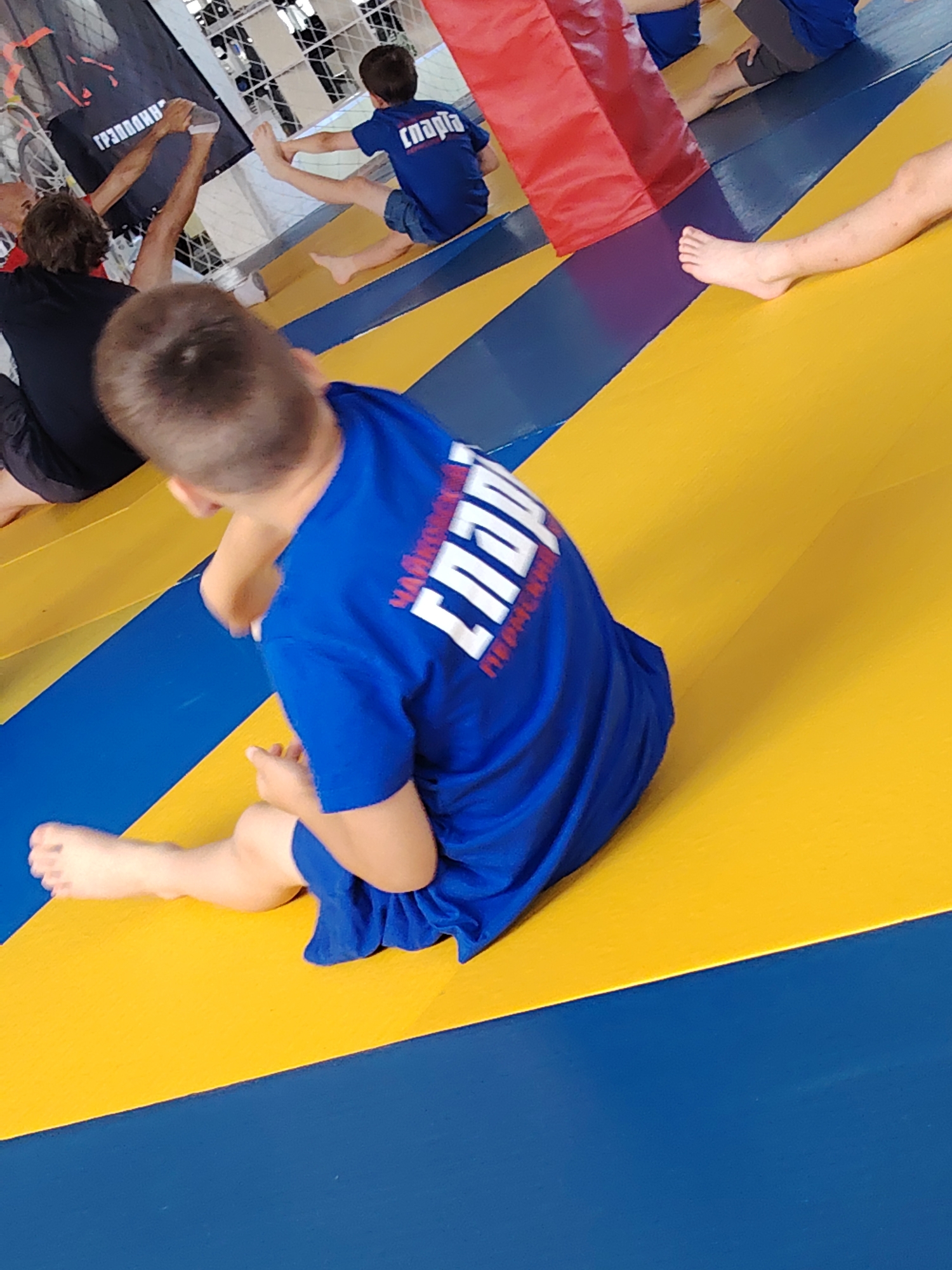 A group of children and adults are engaged in some form of athletic training on a blue and yellow mat. The mat is divided into sections by colored lines. In the foreground, a young boy in a blue shirt with white lettering is sitting, facing away from the camera, with his legs extended. The lettering on his shirt appears to be in Russian and says "СПАРТА" which means "SPARTA." Behind him, another child in a similar blue shirt is also sitting. To the left, an adult's legs are visible, reaching towards a black banner with Russian lettering. To the right, the leg of another person is visible, resting on the mat. The environment appears to be an indoor sports facility. The image captures a dynamic moment during a training session.