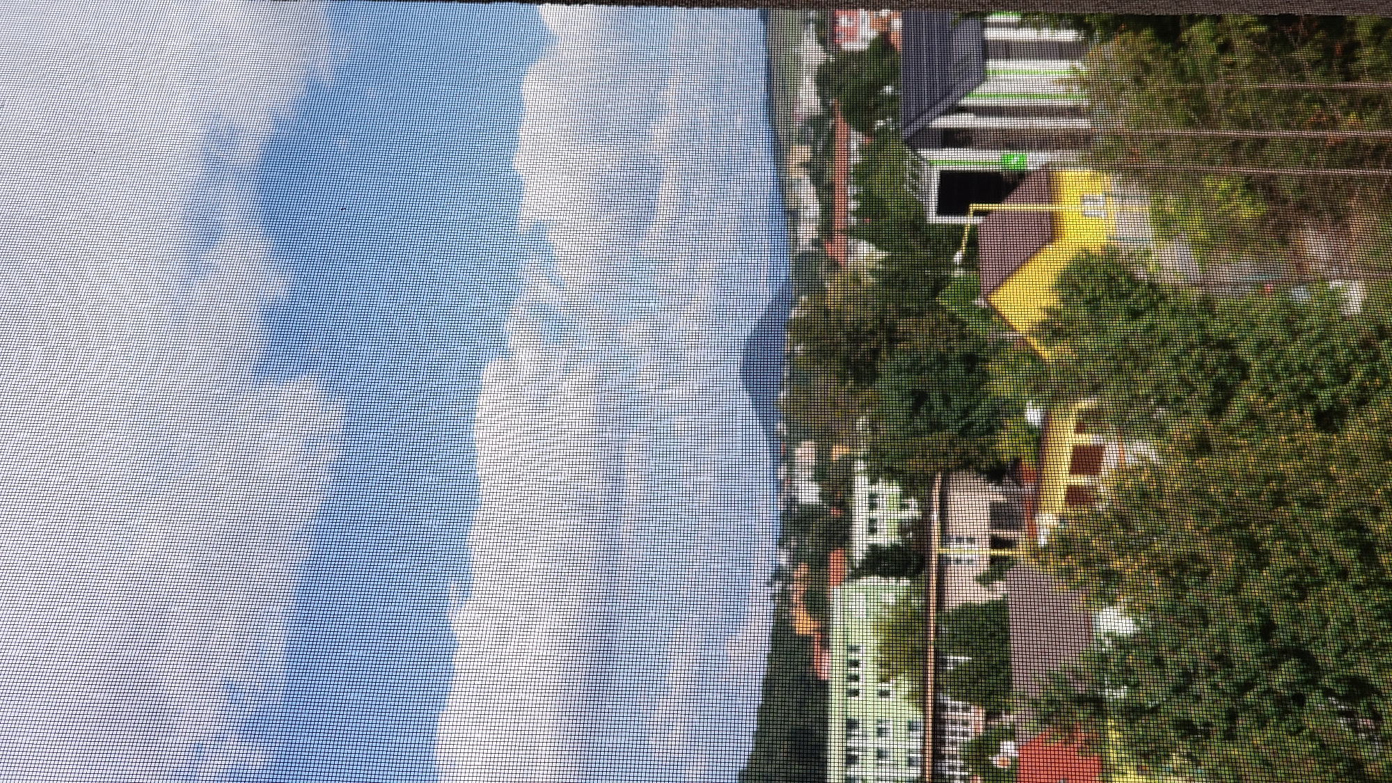 The image depicts a view of the city of Prešov, Slovakia, seen through a window screen. In the foreground, lush green trees are abundant, obscuring much of the lower portion of the scene. Behind the trees, various buildings are visible, including a prominent light green building with multiple windows and a lighter colored roof. To the right of this building, a white structure with an arched entrance is present. Further back, a bright yellow house with a dark brown roof stands out. Adjacent to the yellow house is a building with a dark roof and a distinctive green and white striped facade. In the middle ground, more residential buildings with red and grey roofs are scattered amongst the trees. In the background, a prominent hill or mountain with a conical peak rises above the cityscape. The sky is a vibrant blue, populated by large, fluffy white clouds. The overall impression is a residential area within Prešov, with a natural landscape and urban development integrated.