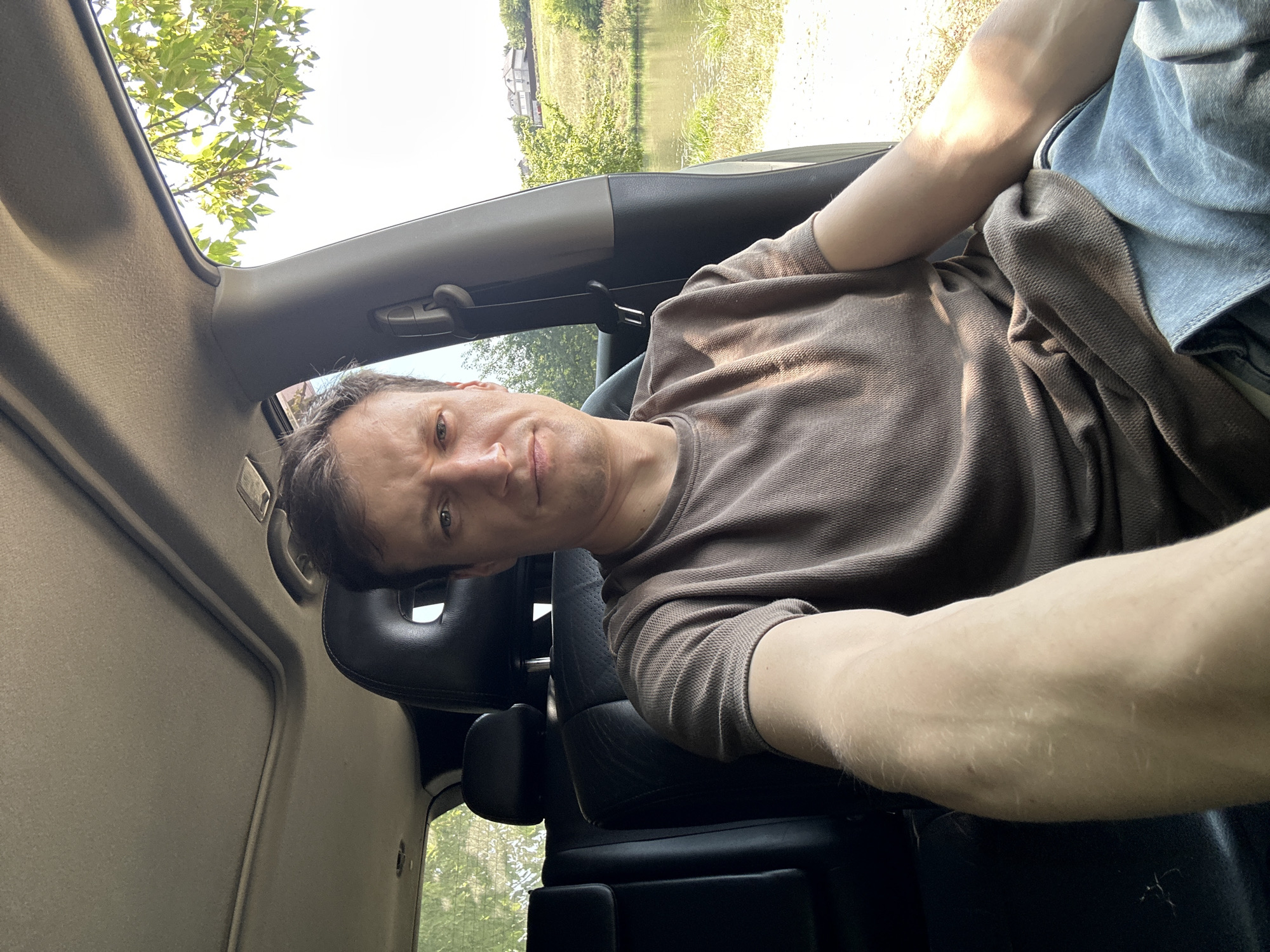 A man is sitting in the driver's seat of a car, with his right arm extended towards the camera. He is wearing a brown t-shirt and blue jeans. The interior of the car is visible, including the headrest of the passenger seat and a portion of the sunroof. Outside the car, a body of water, grassy banks, trees, and houses on a hill are visible. The scene appears to be outdoors in a rural or semi-rural setting, likely during the daytime. The location is specified as Makiv, Ukraine.