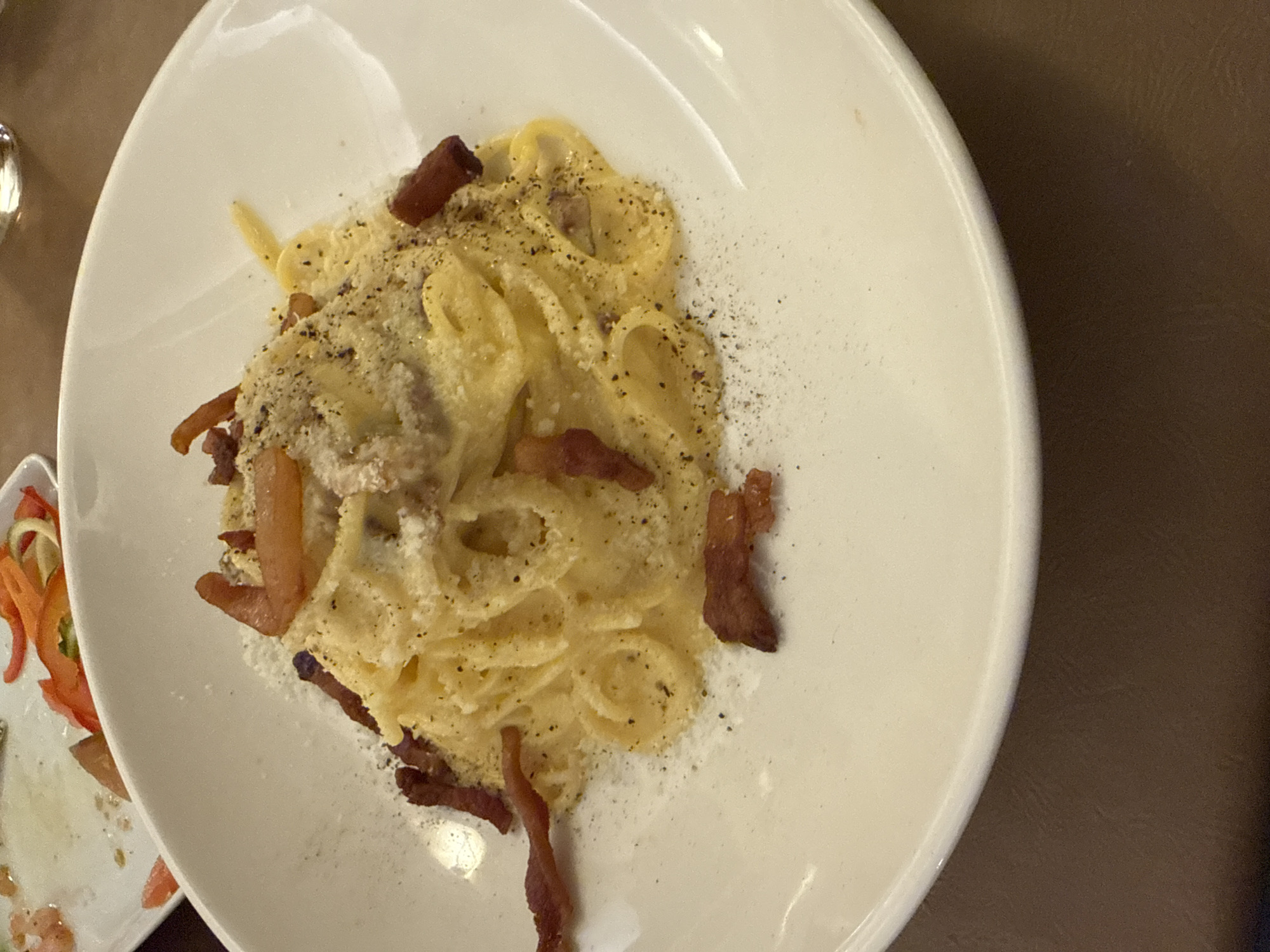 A white, deep, round bowl, centered on a dark brown table, contains a serving of pasta Carbonara, a traditional dish from Rome, Italy. The pasta is coated in a rich, light yellow, creamy sauce, liberally topped with grated white cheese, coarse black pepper, and scattered pieces of dark brown, crispy guanciale. In the upper left background, a partially consumed white plate holds remnants of red and orange bell pepper strips. The setting is an indoor restaurant.