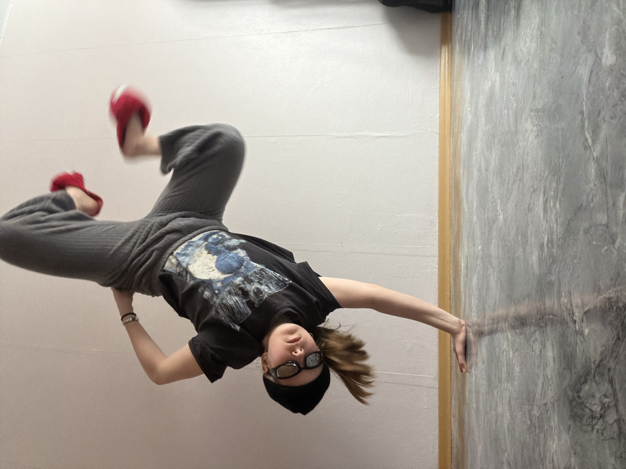 A young person is captured in a dynamic, inverted pose within an indoor setting. They are positioned horizontally across the frame, seemingly defying gravity, with their head towards the right and feet towards the left. Their left hand is extended and braced against a grey, textured wall that resembles marble or concrete. Their legs, clad in red fuzzy slippers, show motion blur, indicating movement, and are angled upwards towards a plain white wall or ceiling.
The individual is wearing a black t-shirt with a prominent blue and white graphic design, possibly depicting a moon or space theme. They also wear grey baggy sweatpants, a black beanie hat, and dark sunglasses. Their hair is swept downwards towards their head due to the inverted pose. The background consists of the aforementioned plain white surface and the grey textured wall, separated by a thin wooden trim. The person maintains a calm expression, looking directly at the viewer. There is no visible text in the image. The time of day is not discernible, but the setting is clearly indoors. No specific location cues are visible within the frame, but the context provided places the event in Perm, Russia.