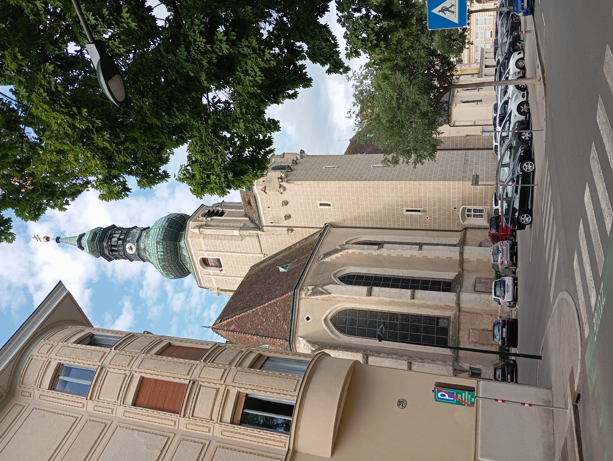 The image shows a street scene in Baden, Austria, featuring a tall church tower with a green, onion-shaped spire and a clock face. To the left of the church is a curved building with ornate facade details and shuttered windows. A traffic light pole with a street lamp extends from the right side of the image, partially obscured by dense green foliage. In the foreground, a street with a pedestrian crossing is visible, lined with parked cars. A blue and white pedestrian crossing sign is also present. The sky is partly cloudy.