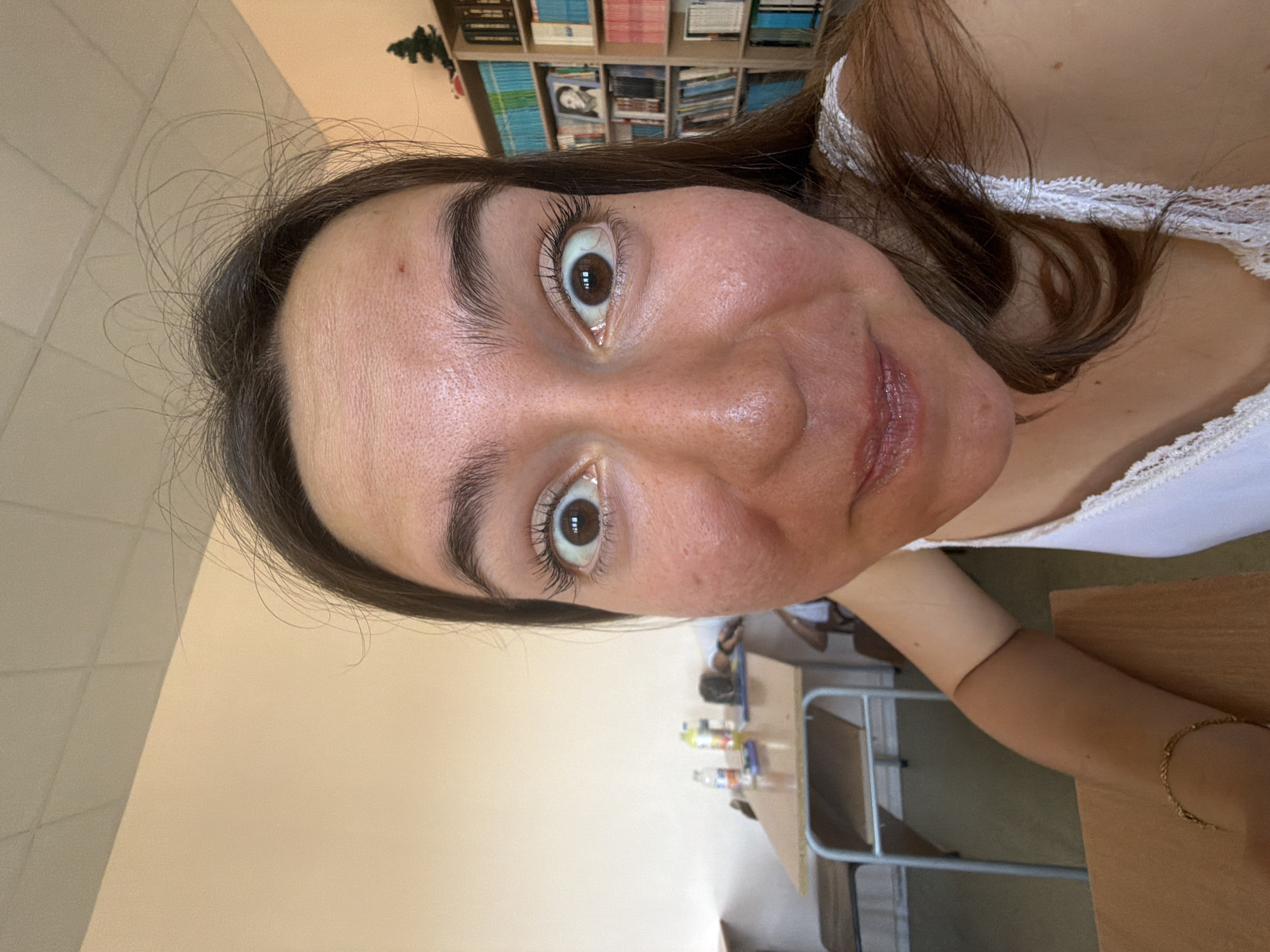 The image is a close-up, distorted selfie taken indoors, likely in a classroom or library setting, as indicated by the bookshelves and desks in the background. The subject is a young woman with dark hair and wide, expressive eyes. Her face dominates the foreground, with a fisheye lens effect creating a dramatic distortion. She is wearing a white top with lace trim. In the background, a desk with water bottles and a cell phone is visible, along with a bookshelf filled with books. The environment appears to be well-lit, with fluorescent lighting on the ceiling. The exact location in Burgas, Bulgaria, cannot be determined from the image alone, as there are no specific landmarks or identifying features.