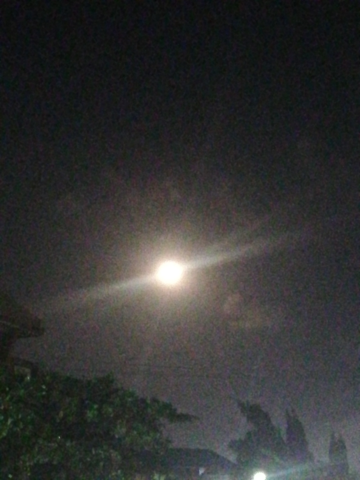 The image depicts a night scene in Lagos, Nigeria. The sky is dark and cloudy with a bright light source, likely a streetlight or the moon, visible through the haze. The foreground contains the silhouetted shapes of trees and rooftops. The location is likely a residential area within the city, but specific landmarks are not discernible.