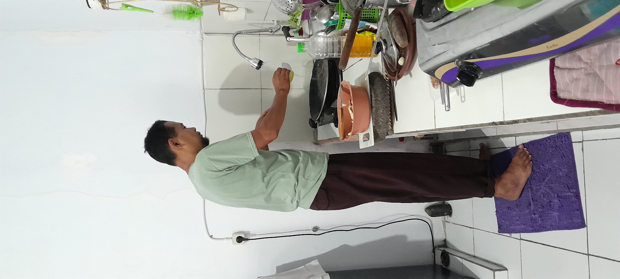 A man is standing in a kitchen, at a sink, washing his hands. The man is wearing a light green t-shirt and dark brown pants. He is barefoot, with his right foot on a purple bath mat. The kitchen has white tiled walls and floors. There is a faucet with running water, a black stovetop, and various kitchen utensils and appliances visible. The scene appears to be in a home in Bendungan, Indonesia.