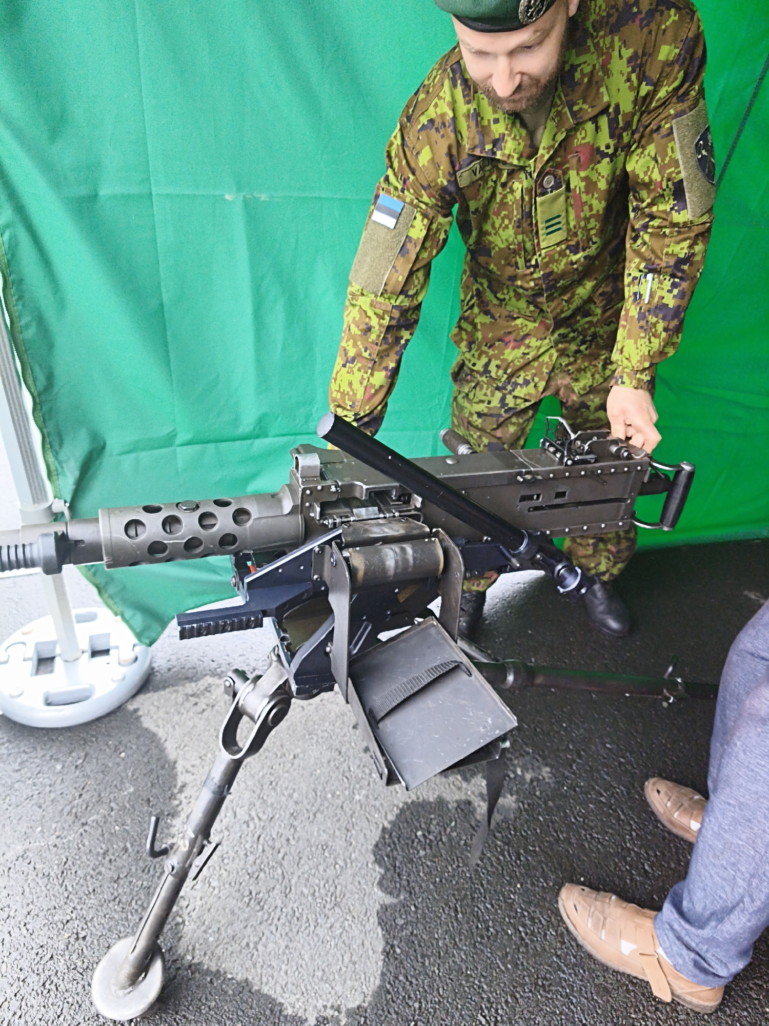 The image shows a man in a camouflage uniform and a green beret, with an Estonian flag patch on his left arm, interacting with a mounted machine gun on a tripod. The man appears to be demonstrating or explaining the weapon, as his hand is on its mechanism. The machine gun is positioned in the foreground and is the primary subject. The background consists of a bright green fabric, likely a backdrop or a tent. The ground is wet asphalt, suggesting it may have recently rained. In the lower right corner, the legs and feet of another person are visible, wearing blue trousers and brown sandals. The scene appears to be an outdoor event or exhibition, possibly in Tallinn, Estonia, given the presence of the Estonian flag.