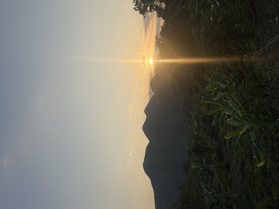 An outdoor landscape photograph captured during sunrise or sunset depicts mountainous terrain in Nha Trang, Vietnam. The sky displays a gradient, transitioning from light blue at the zenith to warm orange and yellow hues near the horizon. The sun, a bright spherical object, is positioned on the mid-right horizon, partially obscured by distant mountain peaks and light clouds. A prominent vertical sun pillar or lens flare extends downwards from the sun, casting a bright golden streak through the lower atmosphere. Multiple mountain peaks, appearing as dark silhouettes, dominate the middle ground against the illuminated sky. The foreground and lower mid-ground consist of dense, dark-green vegetation, with several banana plants identifiable by their broad leaves. The overall scene is backlit, rendering much of the immediate environment in shadow. No individuals, animals, structures, or artificial objects are present.