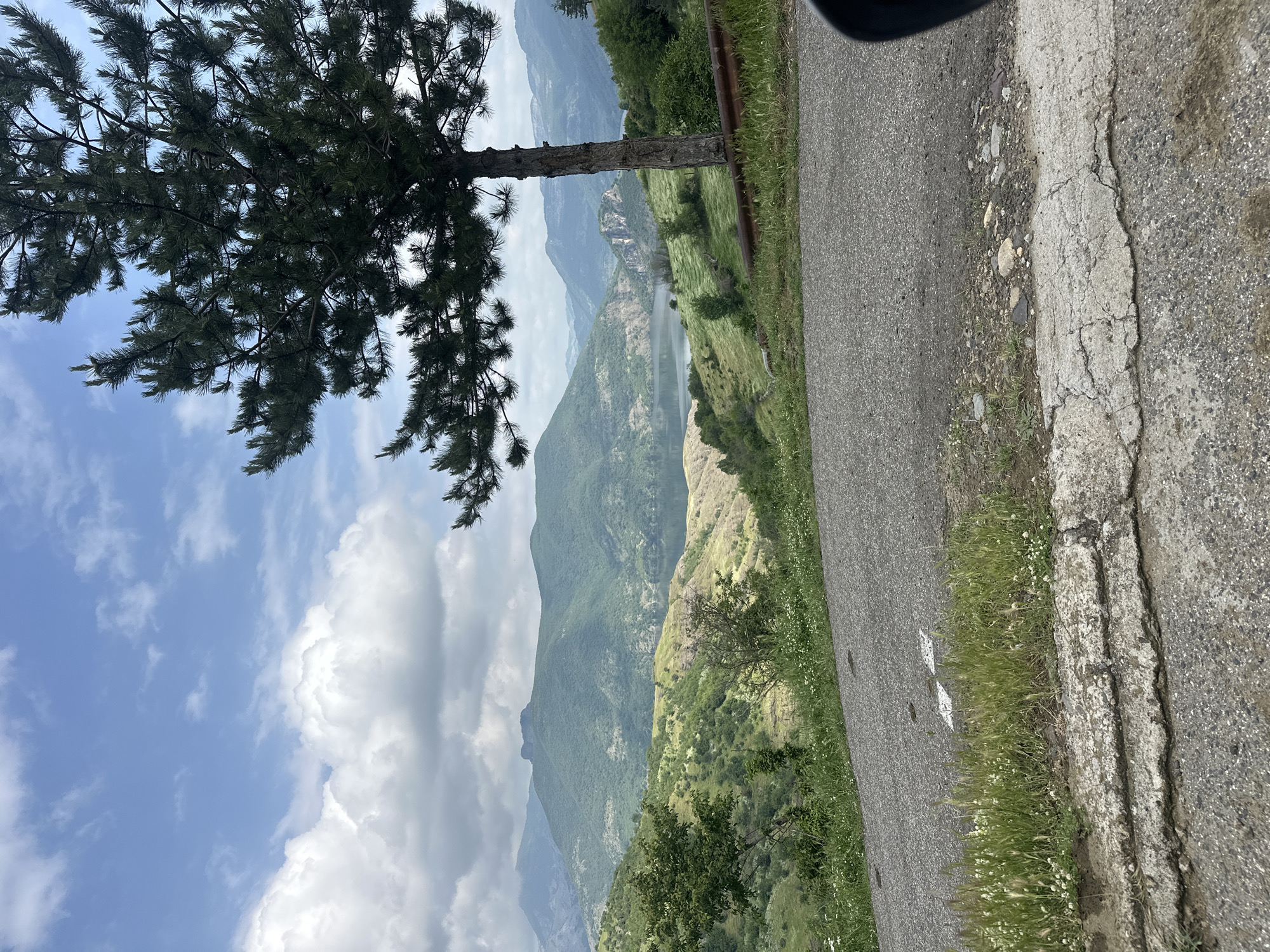 The image displays a picturesque mountainous landscape, appearing as if taken from a vehicle or roadside viewpoint. The original image is rotated 90 degrees counter-clockwise; when viewed correctly, it presents a horizontal landscape.

In the foreground, a gray asphalt road occupies the bottom right, featuring a weathered and cracked concrete shoulder bordered by green grass and small wildflowers. A substantial pine tree, with dark green needles, stands prominently on the left side of the frame, its trunk and branches extending upwards, partially framing the view.

The midground reveals rolling green mountains and hills, densely covered in trees and vegetation, stretching into the distance. A body of water, likely a lake or reservoir, is nestled within the valleys, reflecting the sky and adding depth to the scene. Further back, successive layers of mountains recede, gradually becoming lighter and more subdued in color, suggesting distance and atmospheric haze.

The sky above is partly cloudy, with large, bright white clouds contrasting against a clear blue expanse. The time of day appears to be daytime, likely late morning or early afternoon, given the bright illumination and clear visibility. The weather is fair. There are no people visible, and no specific activities are taking place, the scene captures a serene natural vista. No discernible text is visible, only a faint, faded white road marking on the asphalt. The setting is a natural, mountainous environment, consistent with the provided location context of Star Chitak, Bulgaria.