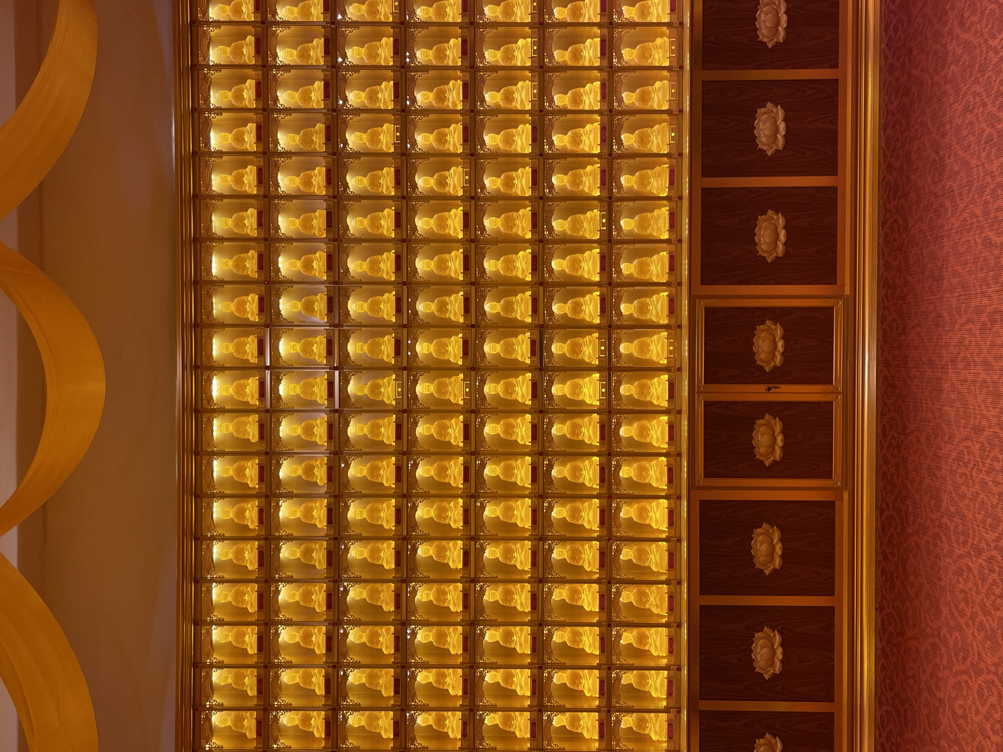 The image presents a section of the interior of a temple, likely located in Yongding District, China. A grid of small, illuminated Buddha statues in individual glass-fronted compartments dominates the central area. To the right of the Buddhas is a section of a dark wood cabinet with lotus flower carvings on each panel. A red patterned wall appears on the far right. To the left of the Buddhas is a curved, golden decorative element on a light-colored wall. The focus is on the illuminated Buddhas, suggesting reverence or commemoration.