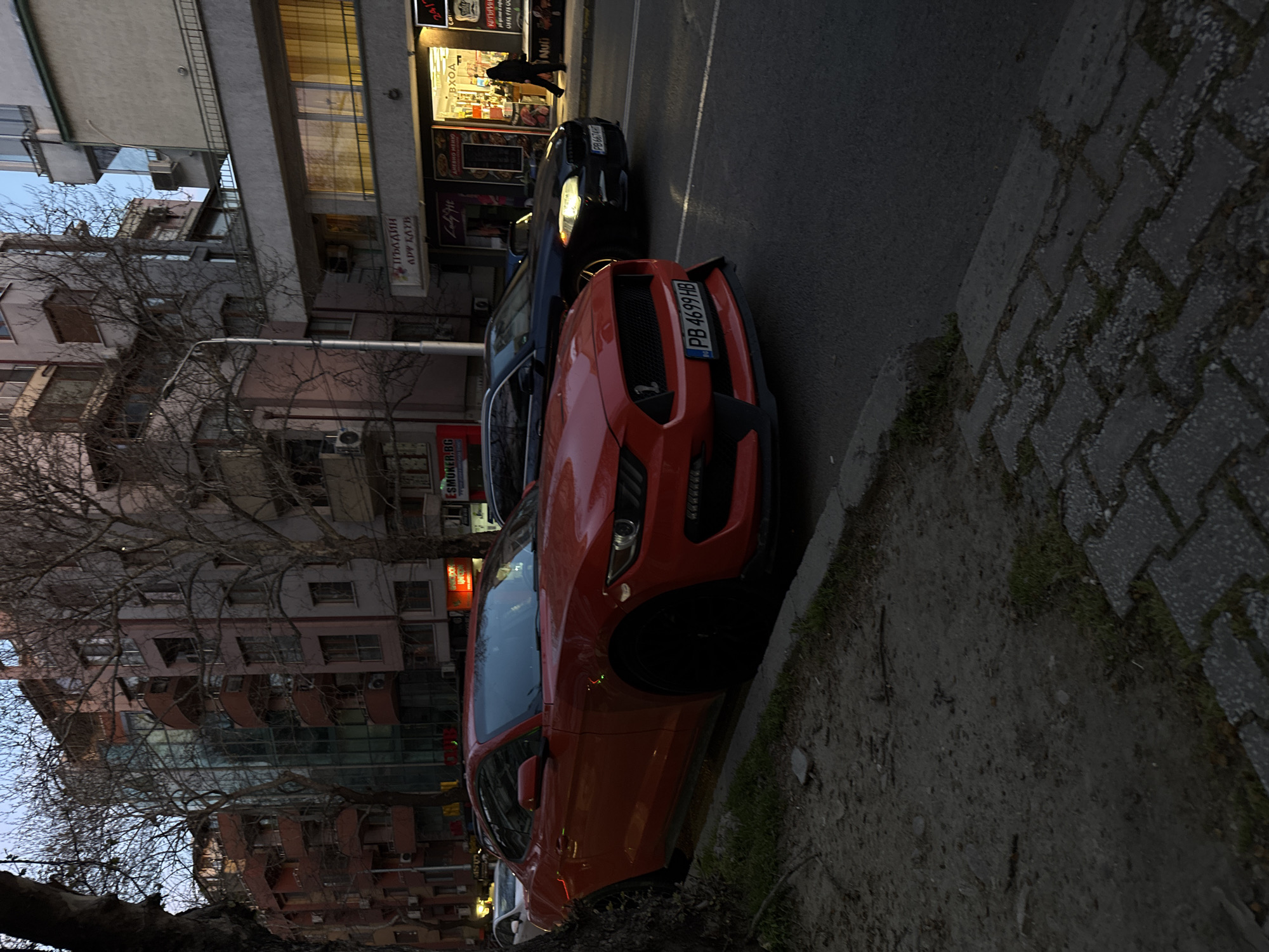 Here is a description of the image.
The image shows an outdoor scene on a street in what appears to be the city of Plovdiv, Bulgaria. Two cars are parked along the curb. The car closest to the viewer is an orange sports car with the license plate "PB 4699 HB." Behind it is a dark-colored car with the license plate "PB 6674 HT."
Behind the cars are buildings, including shops and residential buildings. A sign that reads "ESMOKER.BG" is partially visible on one of the buildings. Another shop sign reads "Lady Fit." There is a person walking in front of one of the shops. Trees with bare branches are also visible. The time of day seems to be late afternoon or early evening, judging by the lighting conditions.
